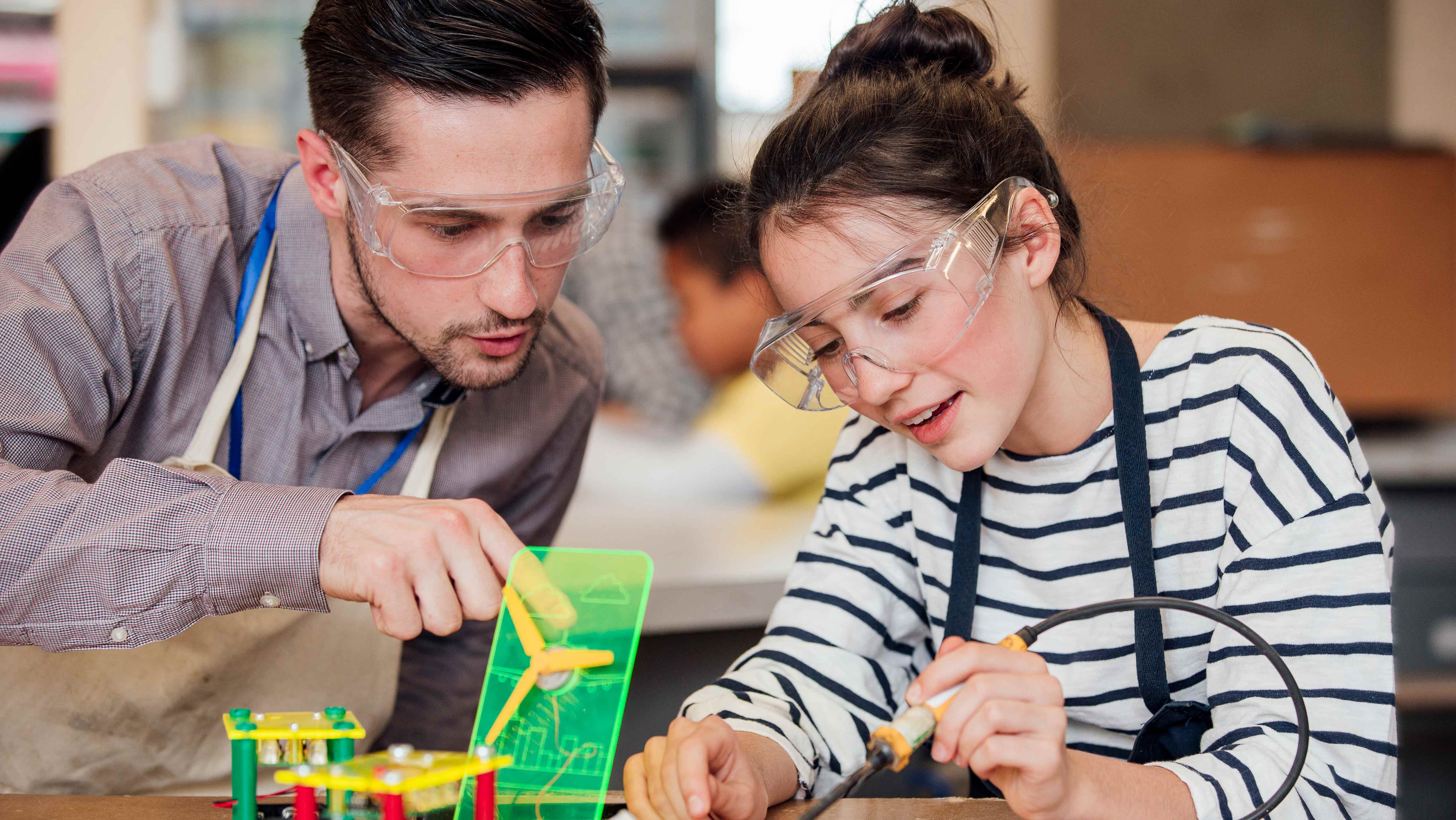 A teacher and a student, both wearing safety glasses, working on a project involving a soldering iron and red, green and yellow components of varying shapes.