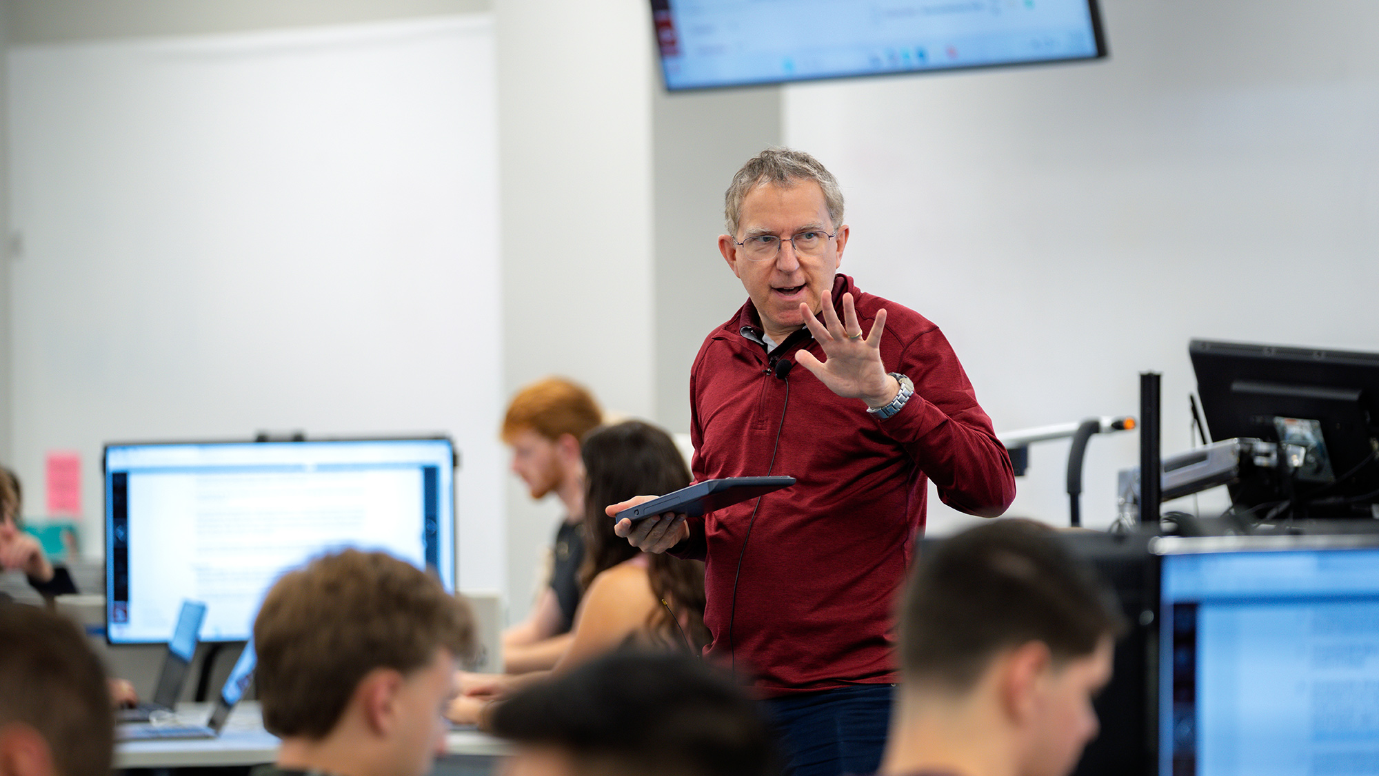 A man holding a keyboard and teaching a room full of students who are sitting at computers.