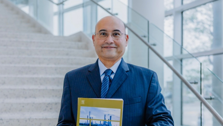 A man wearing a blue suit stands in a modern building, holding a book. 