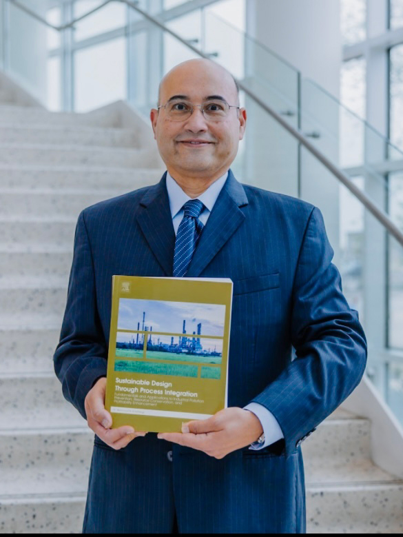 A man wearing a blue suit stands in a modern building, holding a book titled Sustainable Design: Global Perspectives. 