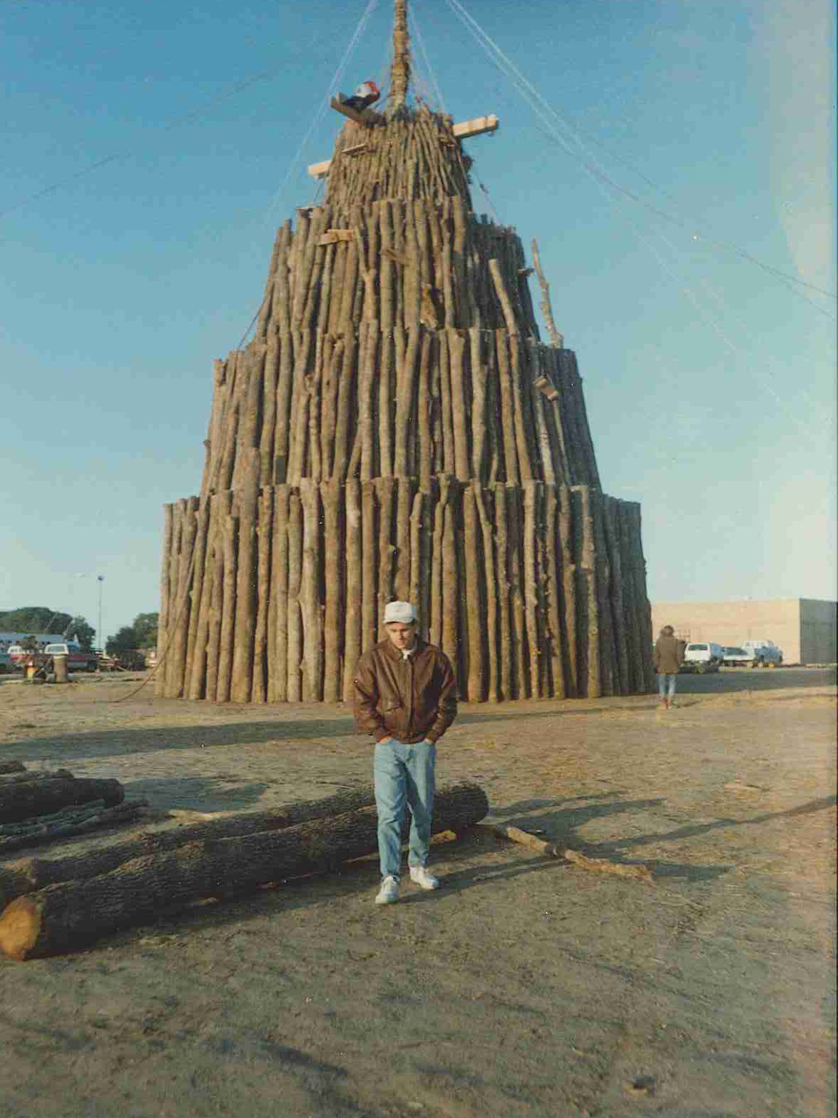 Man stands in front of a large wood pile outdoors.