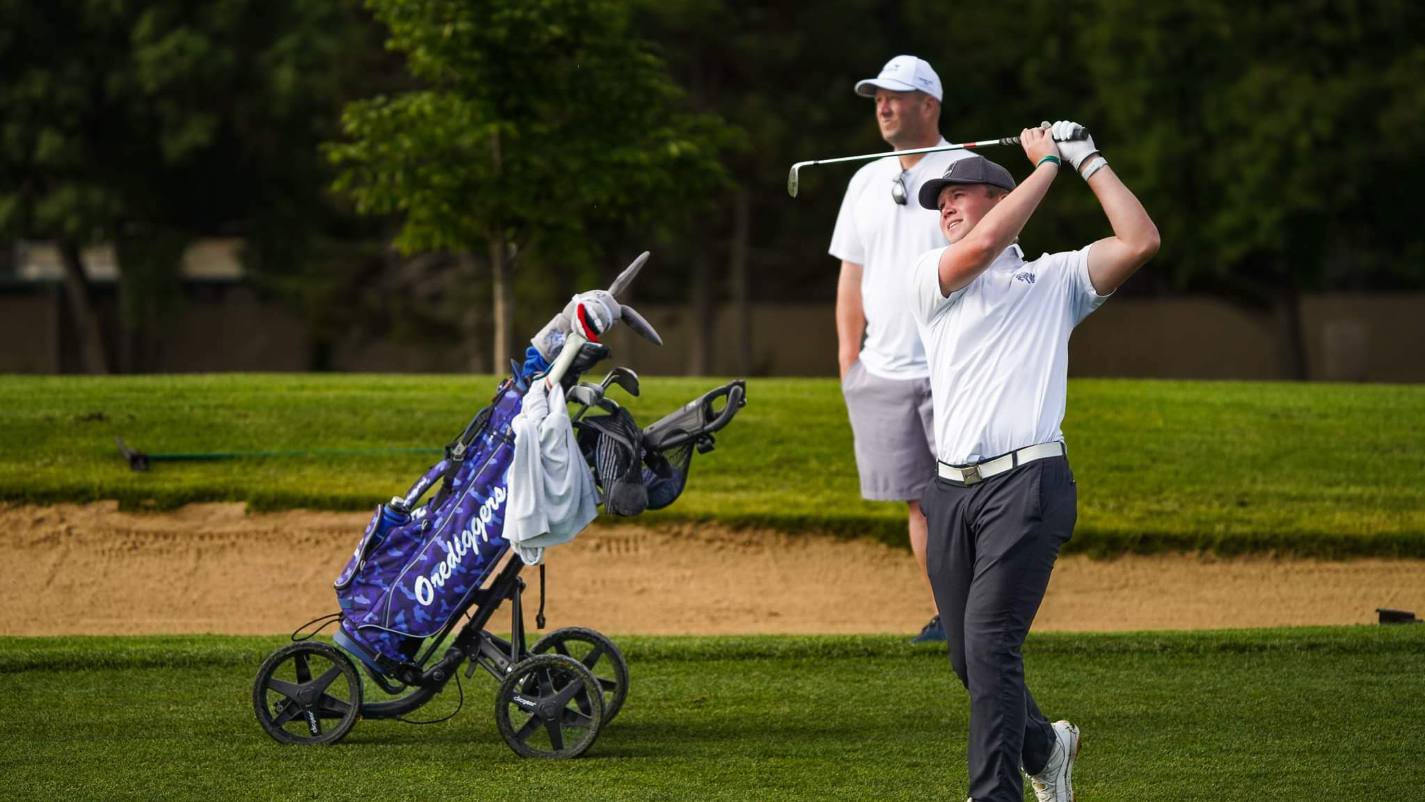 A man on the golf course after swinging a ball, with another man and golf bag in the background.