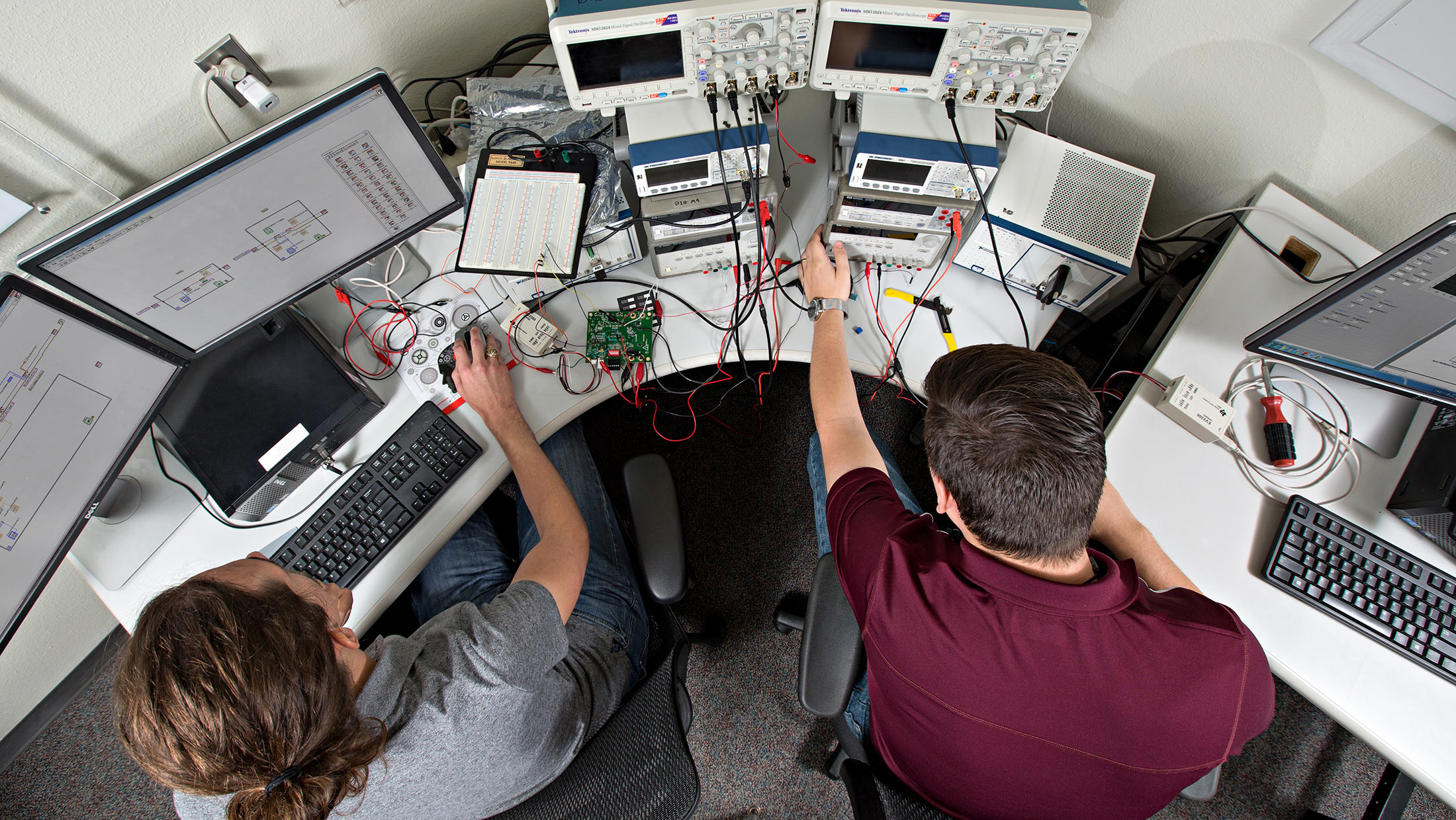 Two individuals are seated at a workstation surrounded by electronic equipment, including multiple monitors and testing devices.