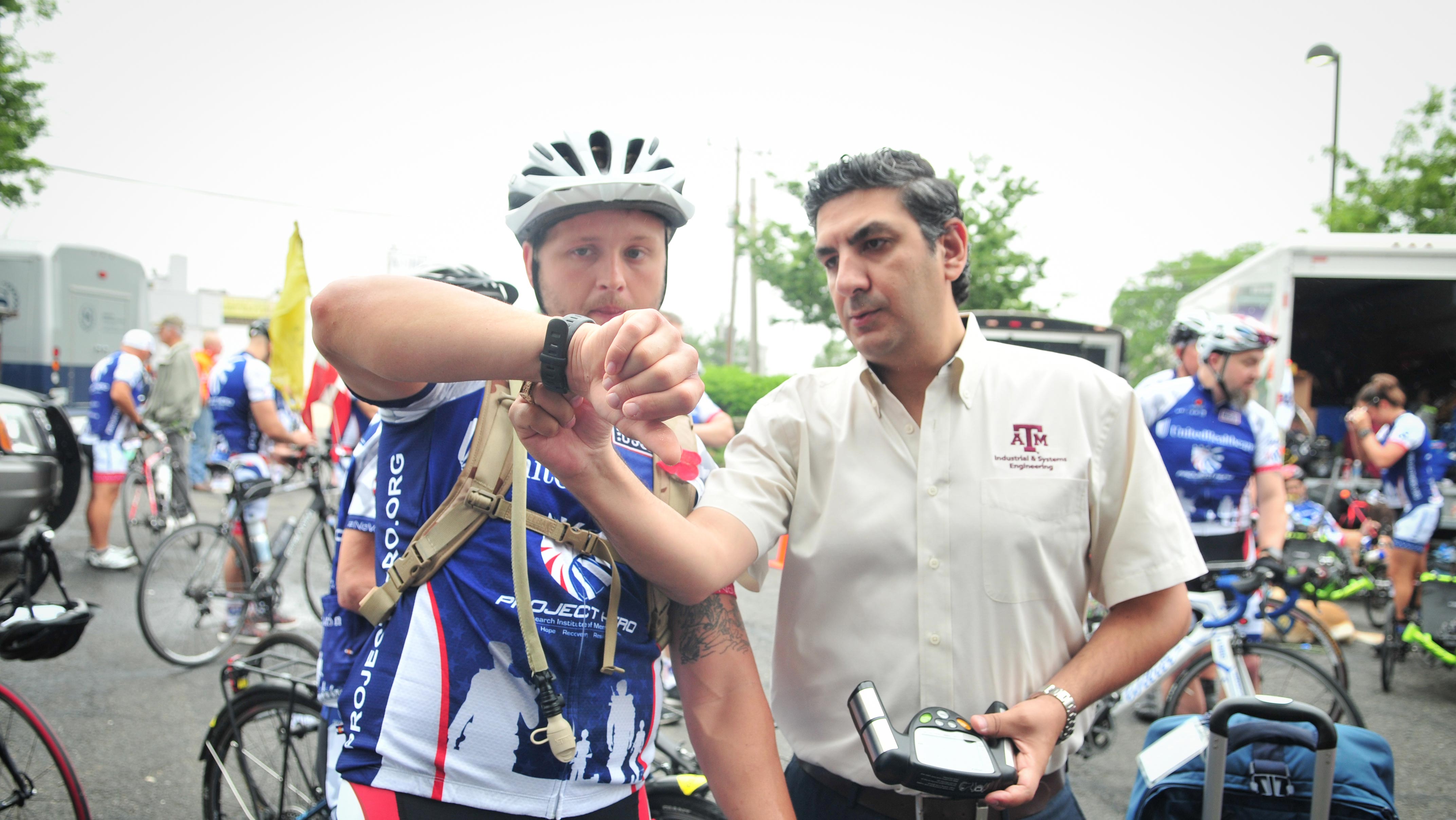 A man wearing a cycling jersey and helmet shows his wrist device to Dr. Sasangohar in a white Texas A&amp;M shirt at a cycling event. Other cyclists and bikes are visible in the background.