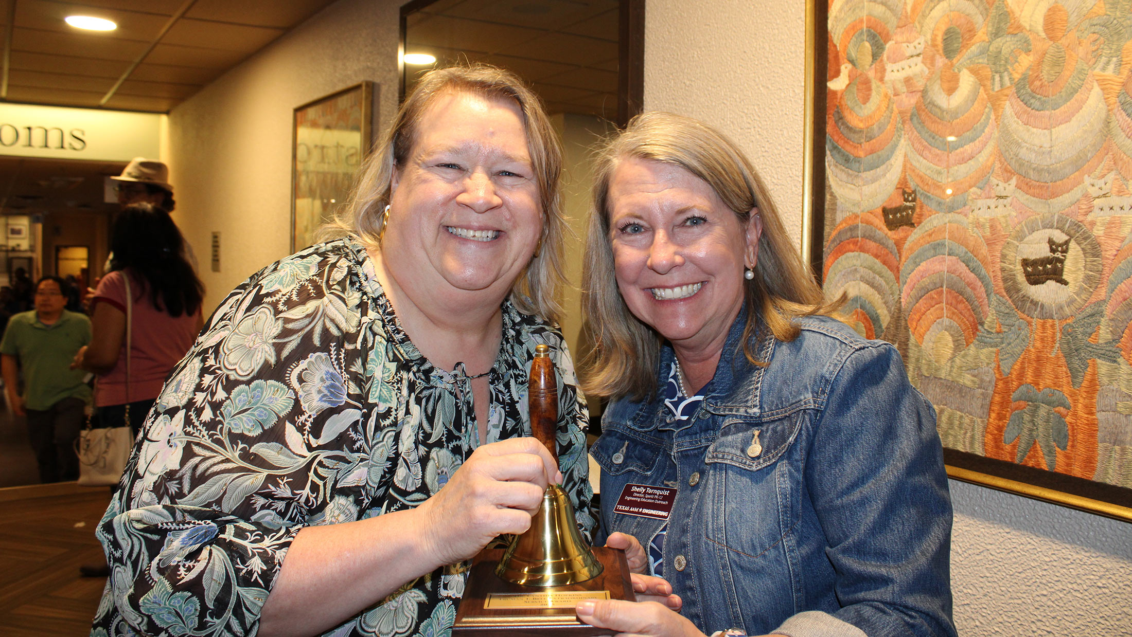 Two women holding a hand bell on a wooden plaque smile at the camera.