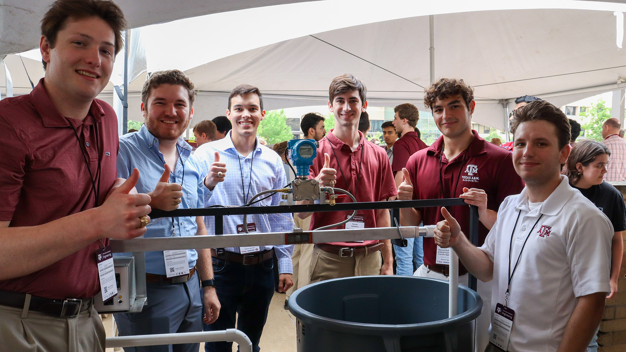 Six people with their thumbs up standing outside under a tent around a trash can with tubes running through it.