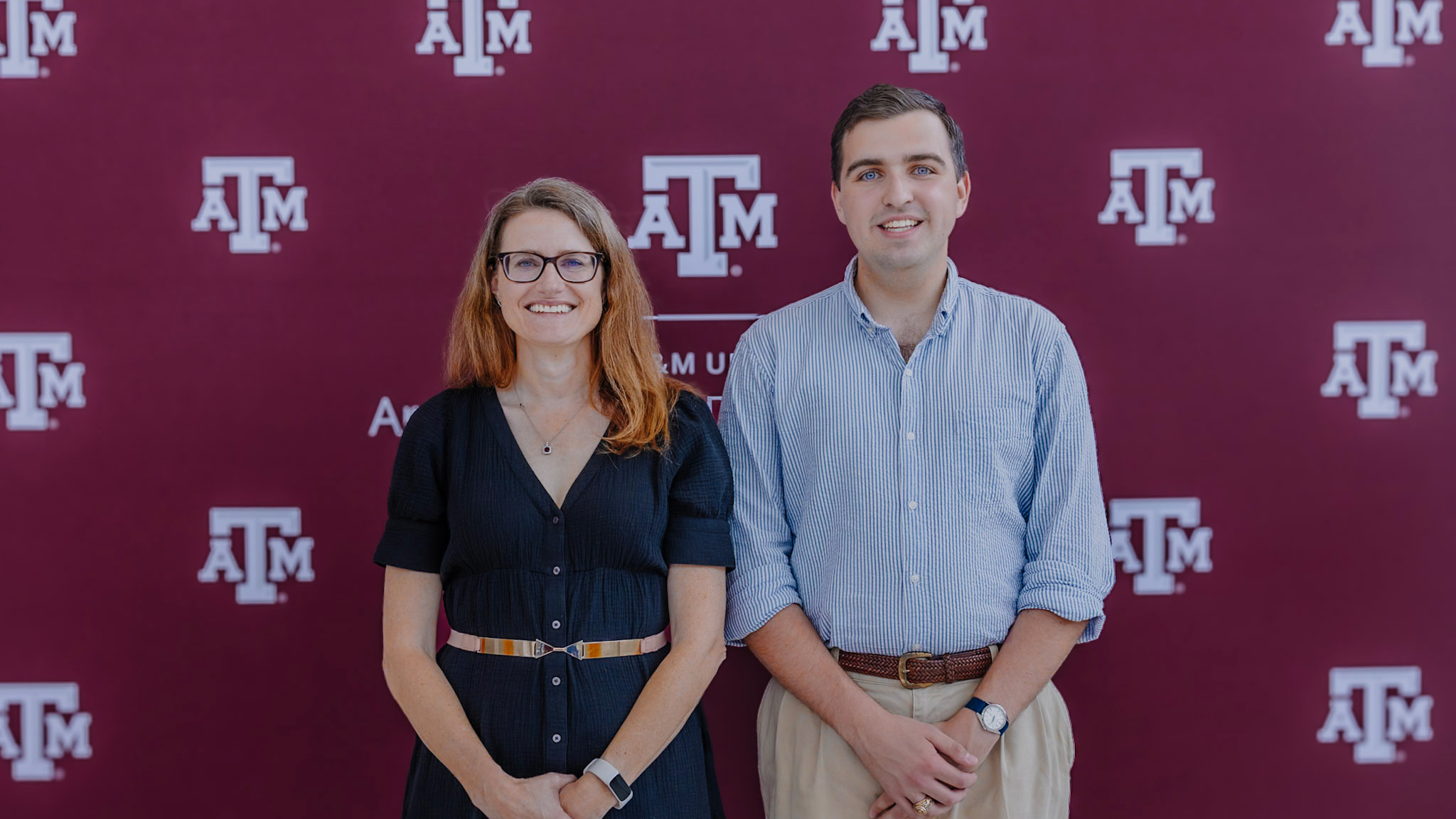 Two individuals stand in front of a maroon backdrop featuring the Texas A&amp;M logo.