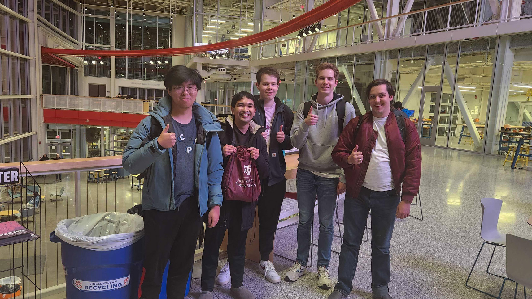 Five students stand smiling and giving thumbs-up gestures in a modern indoor space with large windows and open architecture.  