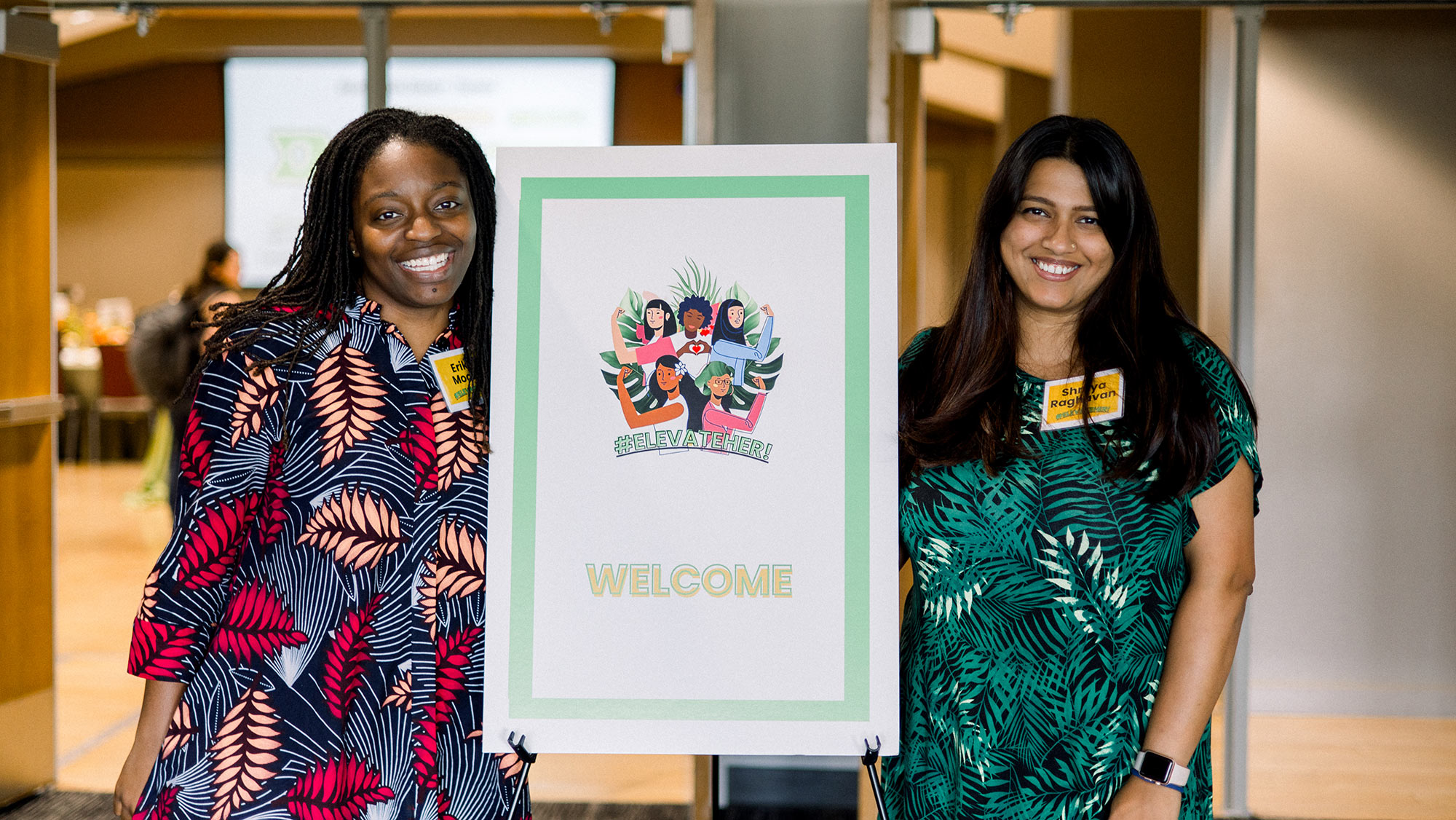 Two women stand next to a welcome sign. 