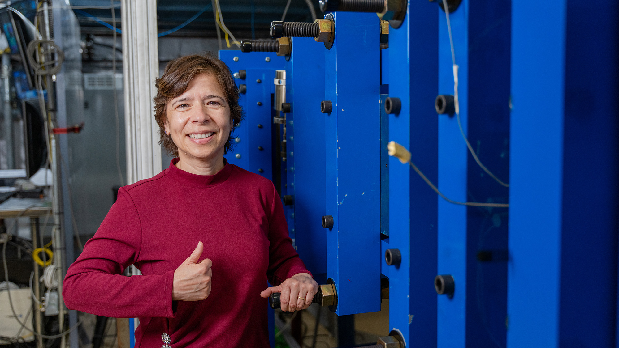 Woman smiling and giving a thumbs-up in a lab with large equipment. 