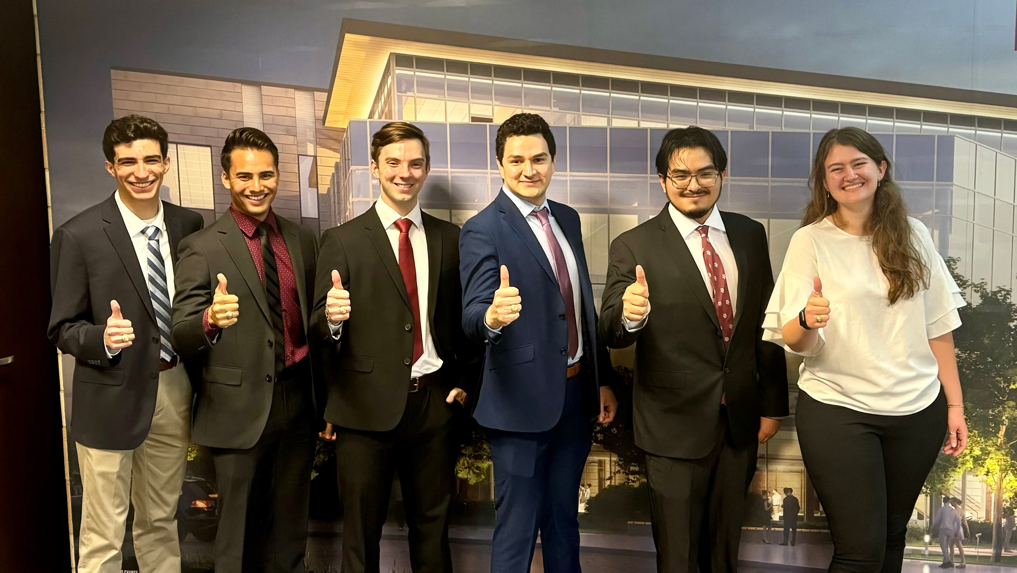 Six individuals in business attire pose in front of a wall graphic with thumbs up.