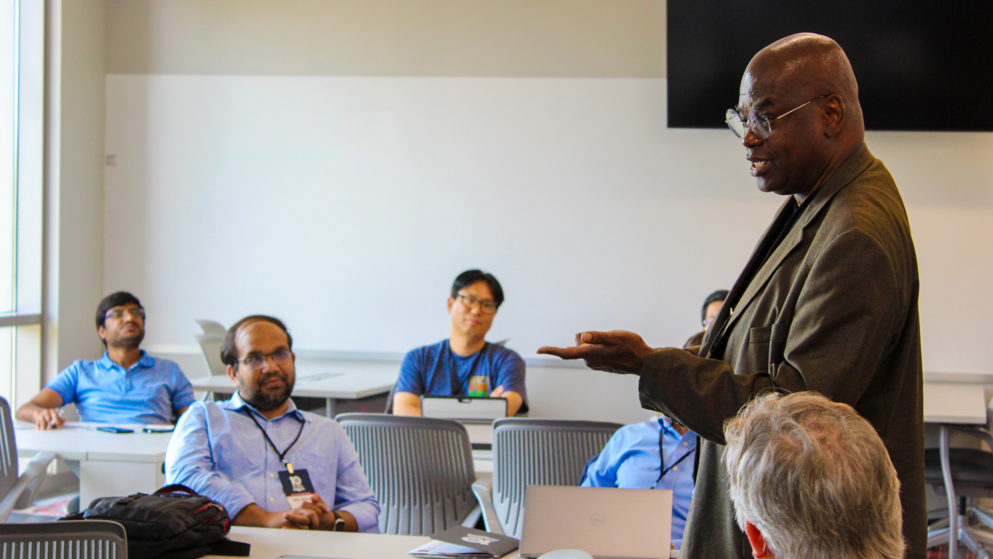 Man stands in front of a desk talking to other adults.