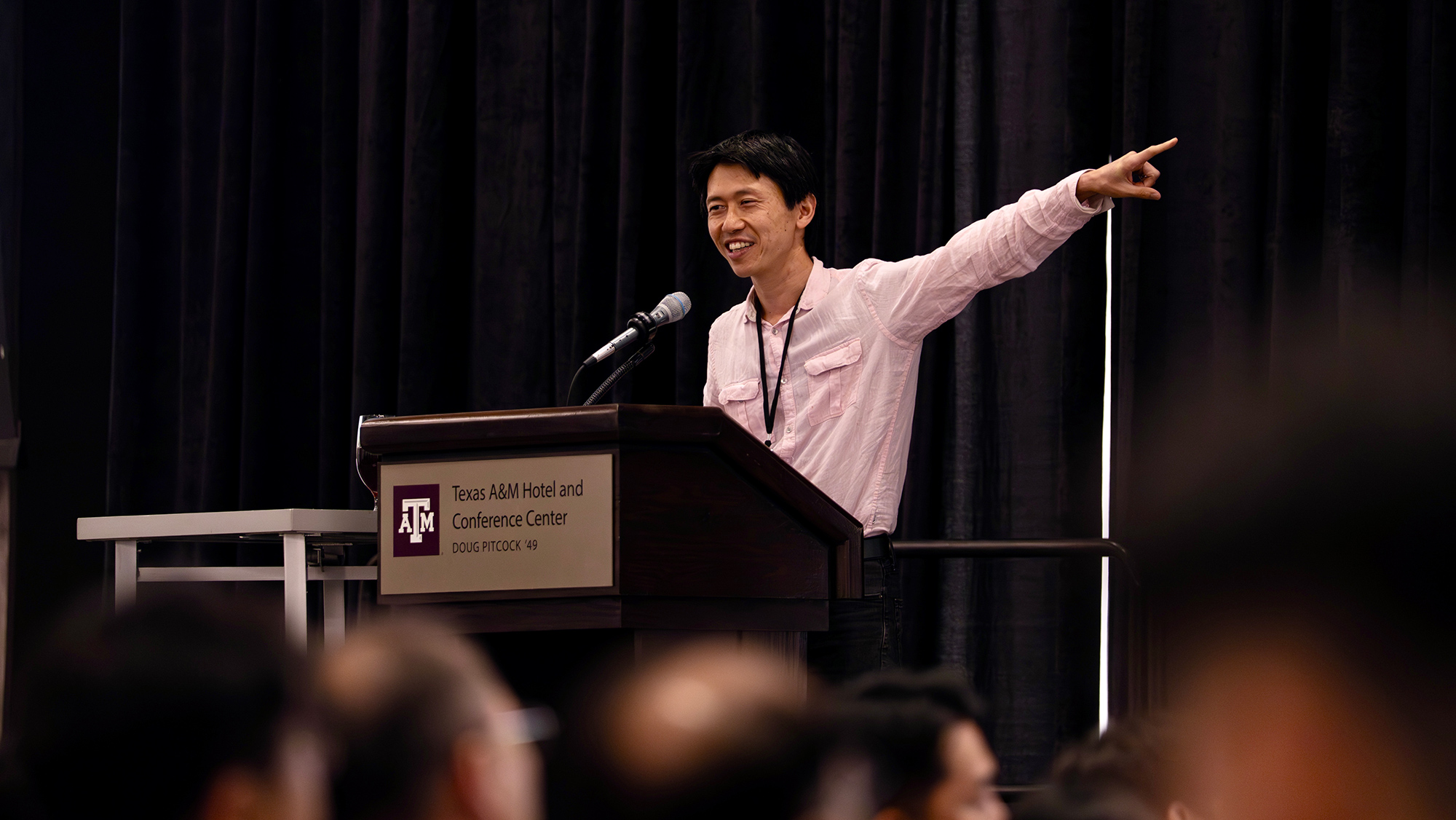 A speaker stands at a podium, pointing and engaging with an audience. 