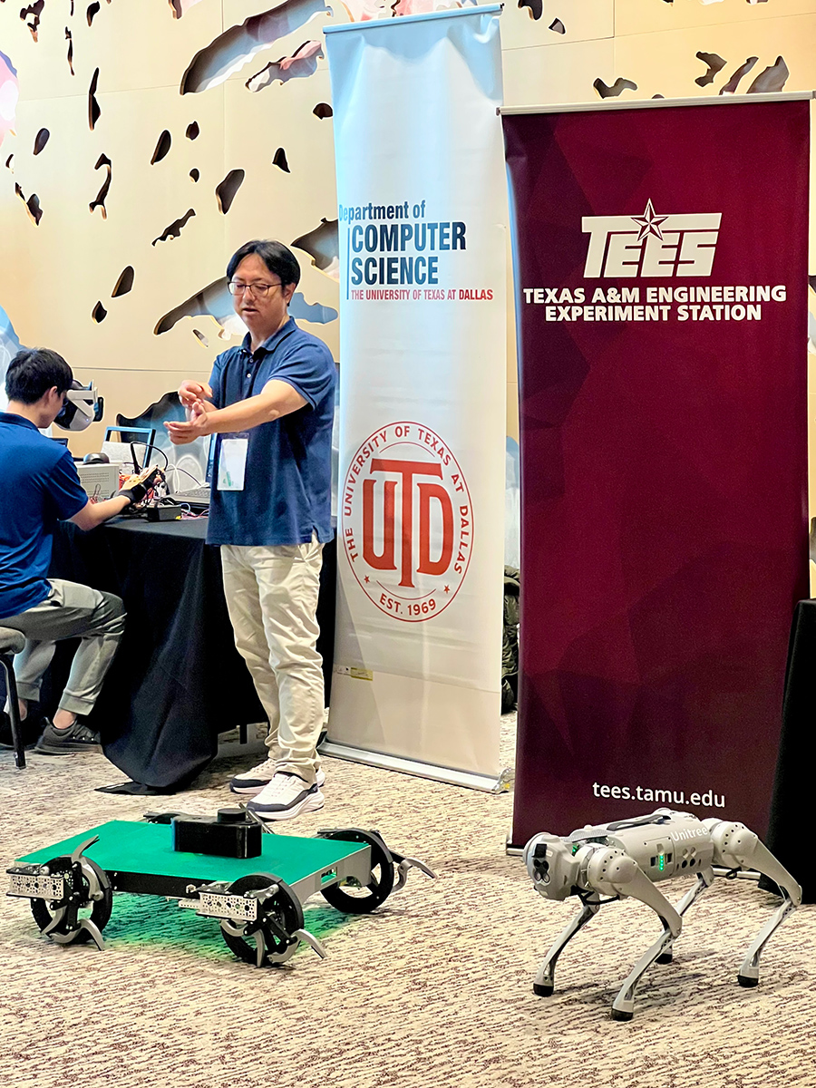 A person stands beside two robots at a display featuring banners for the University of Texas at Dallas and Texas A&amp;M Engineering Experiment Station. 