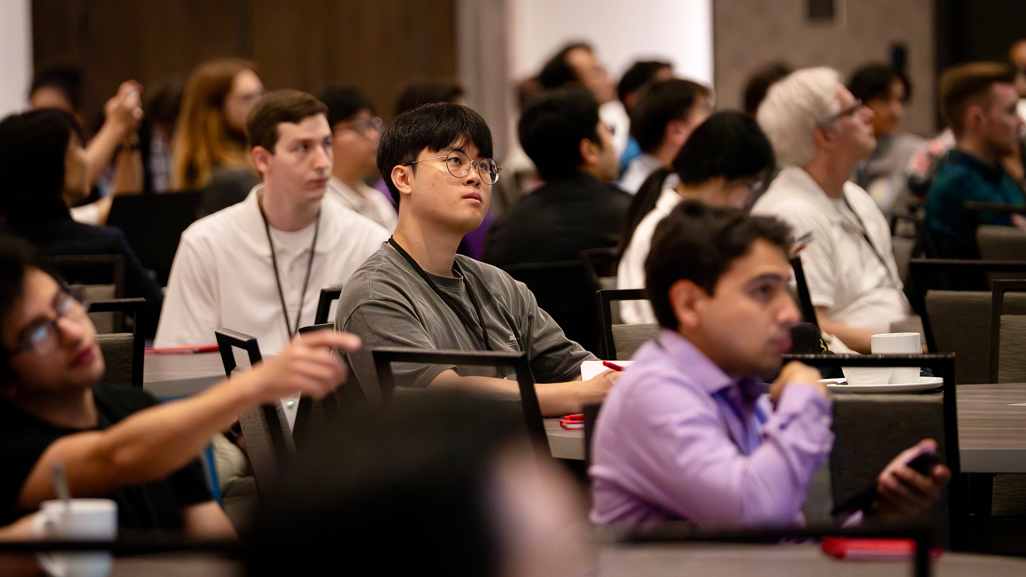 Individuals sit at tables in a conference setting, focused on a speaker. 
