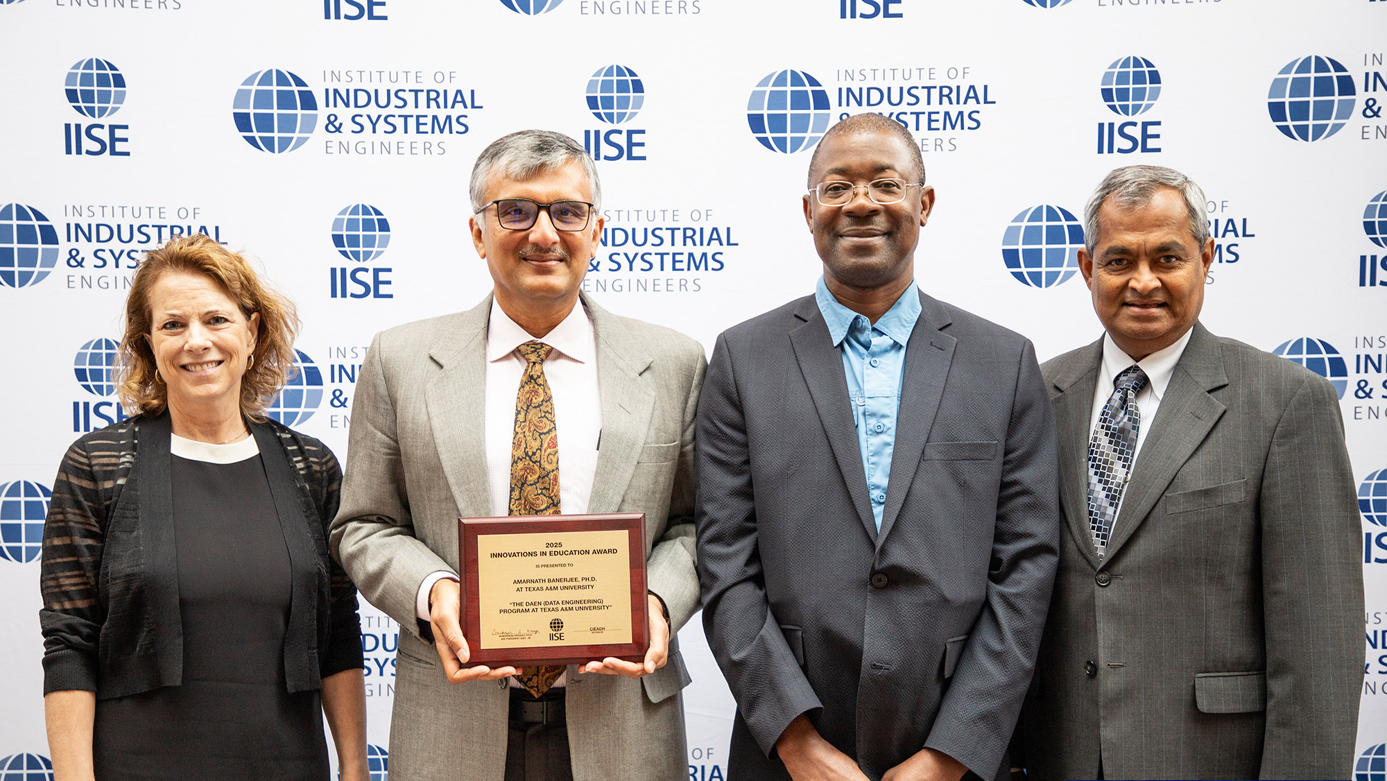 Four individuals stand in front of a step-and-repeat banner with the Institute of Industrial and Systems Engineers logo. The second person from the left holds a plaque. All are dressed in professional attire and smiling for the photo.  