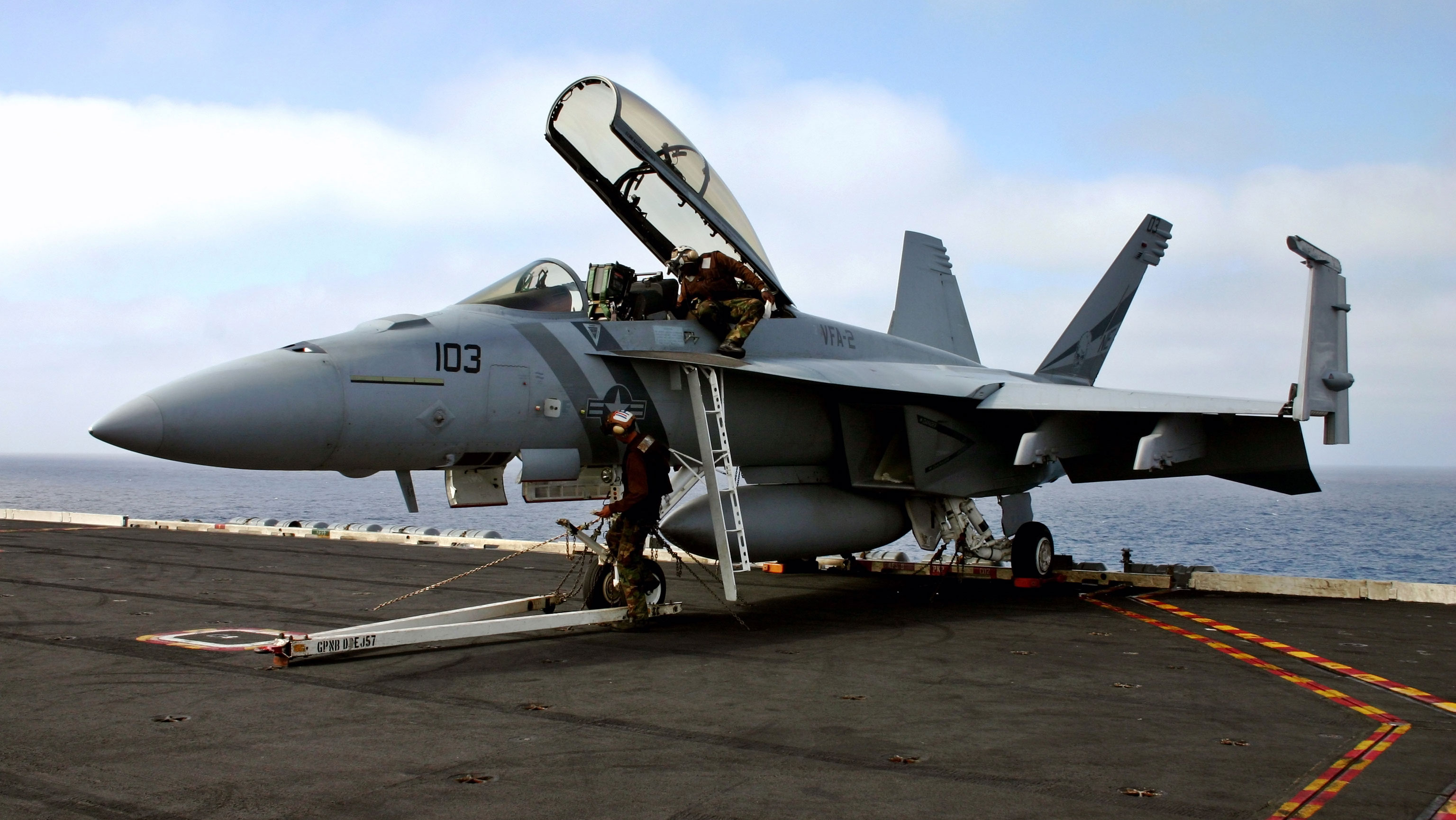 A fighter jet with its wings folded upward and parked on the flight deck of an aircraft carrier at sea. 