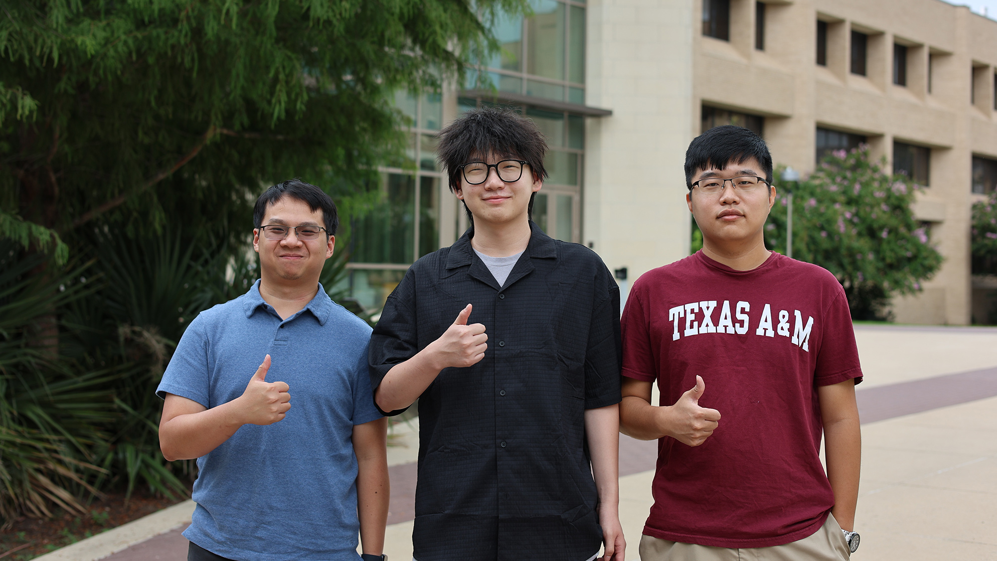Three students standing, smiling and giving a thumbs-up outside of a building.  