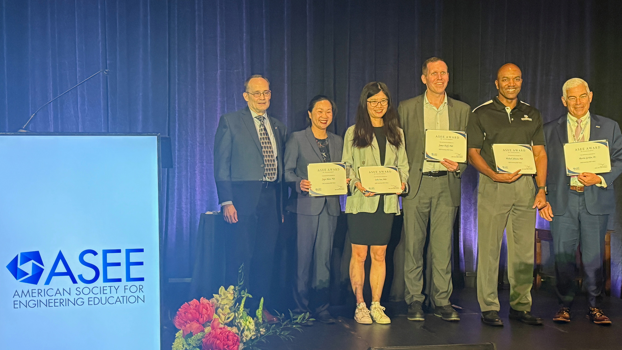 A group of six individuals stands on stage at an event organized by the American Society for Engineering Education. They are holding certificates and smiling.