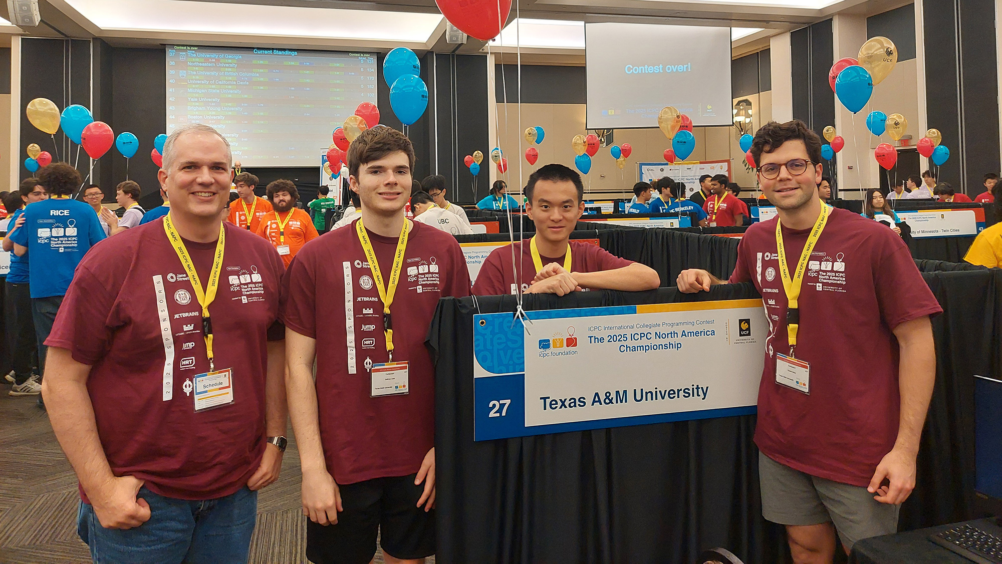 Four men stand in a conference room next to a sign for Texas A&M University that has balloons attached to it.