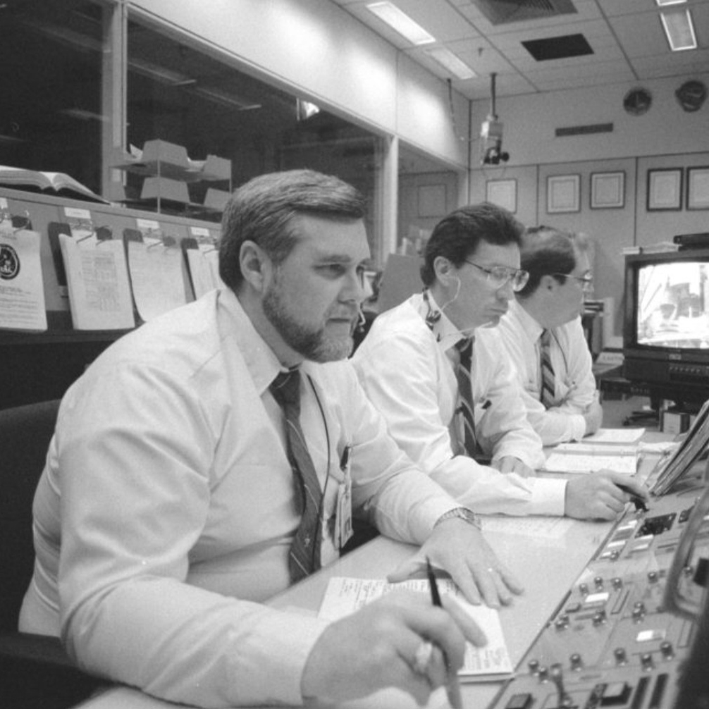 Three men sit at a console in a flight control room. 