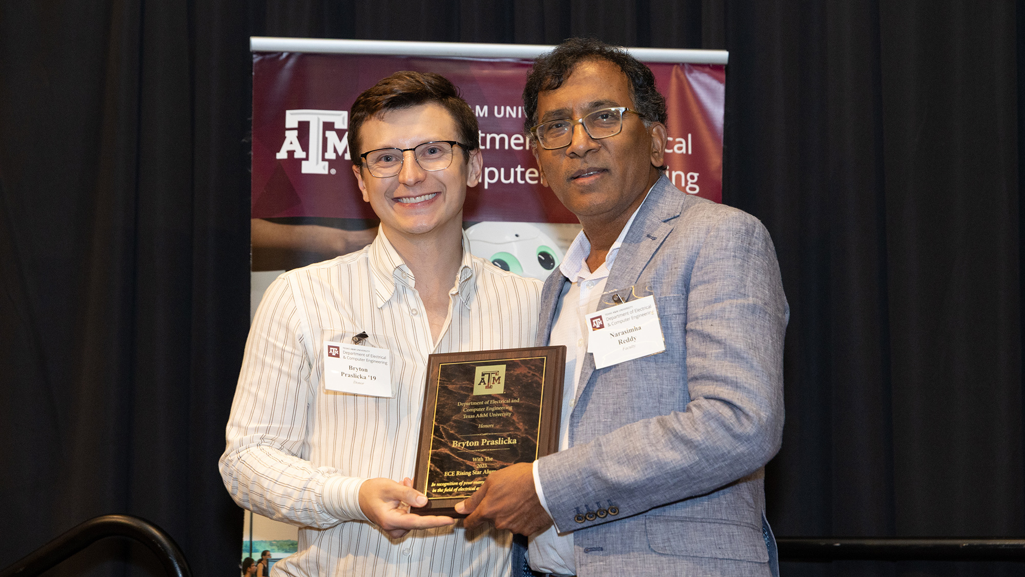 Two men hold an award, smiling and standing in front of black curtains.