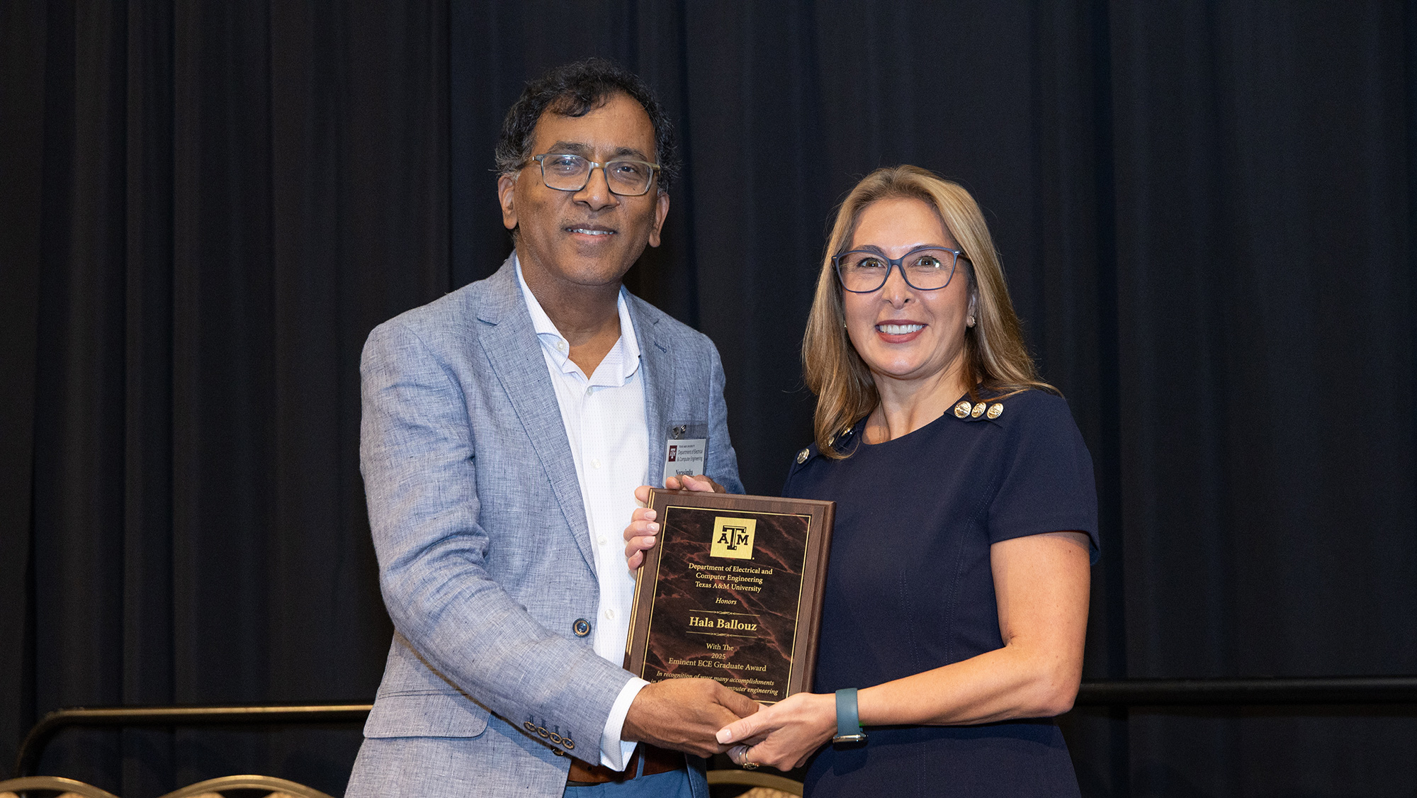 A man and a woman hold an award, smiling and standing in front of black curtains.