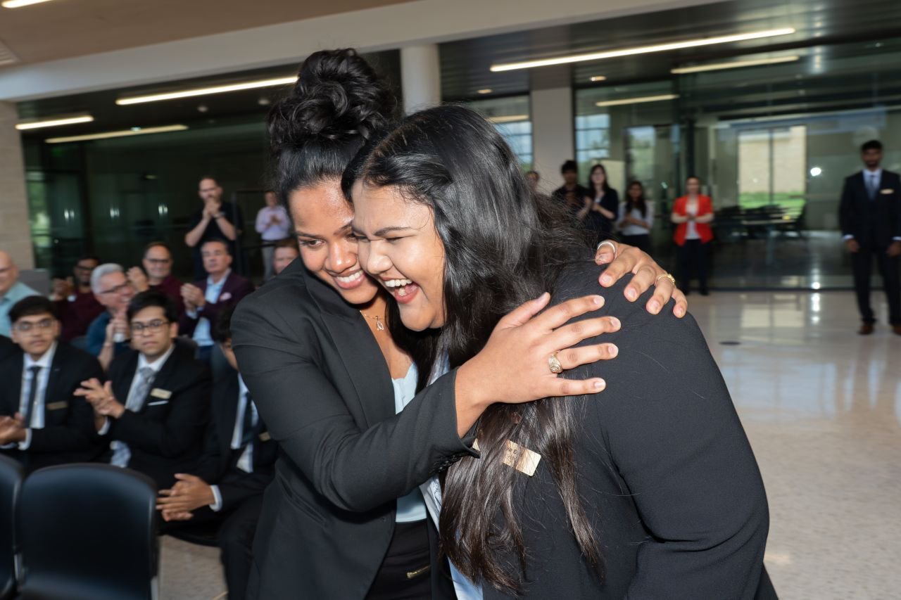 Two individuals in business attire smile and hug.