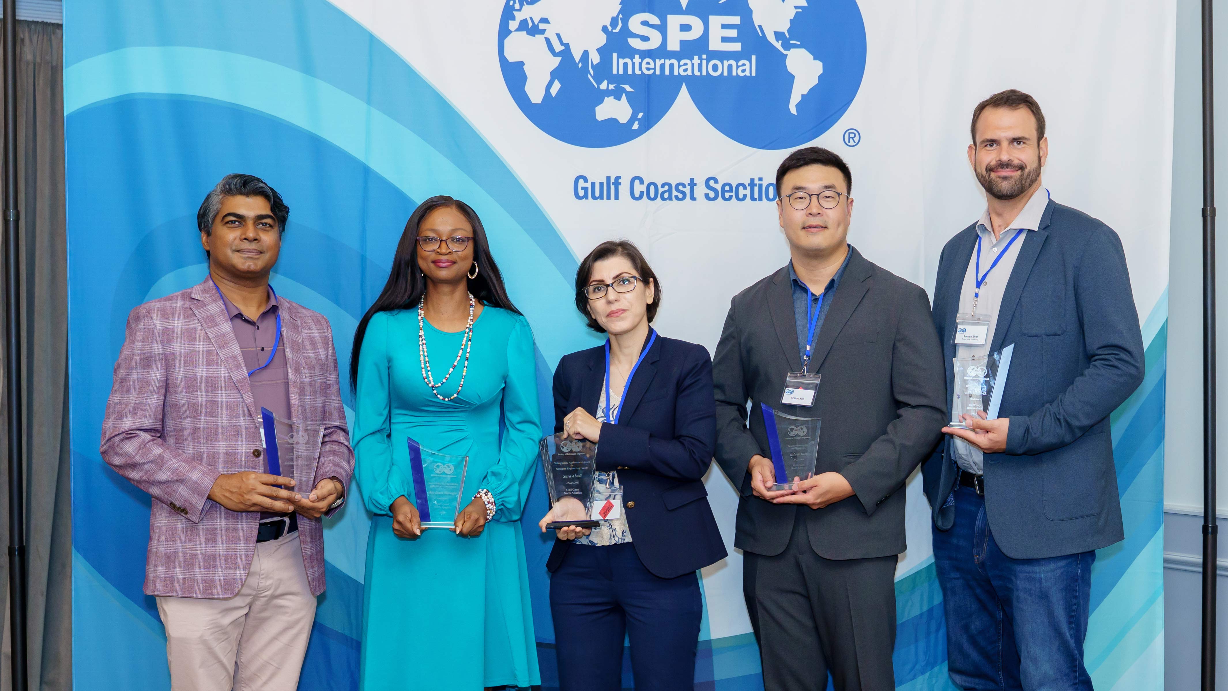 Five individuals stand in front of a backdrop holding awards. They are in professional attire. 