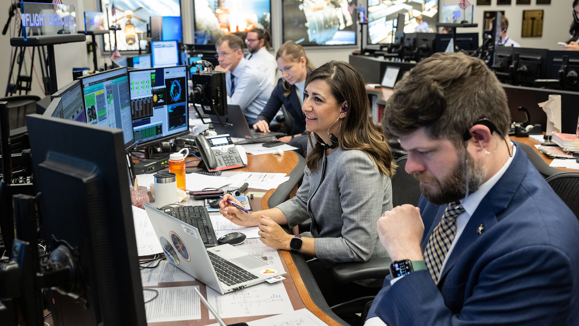 People working at a multimonitor desk with headsets on in a mission control center.