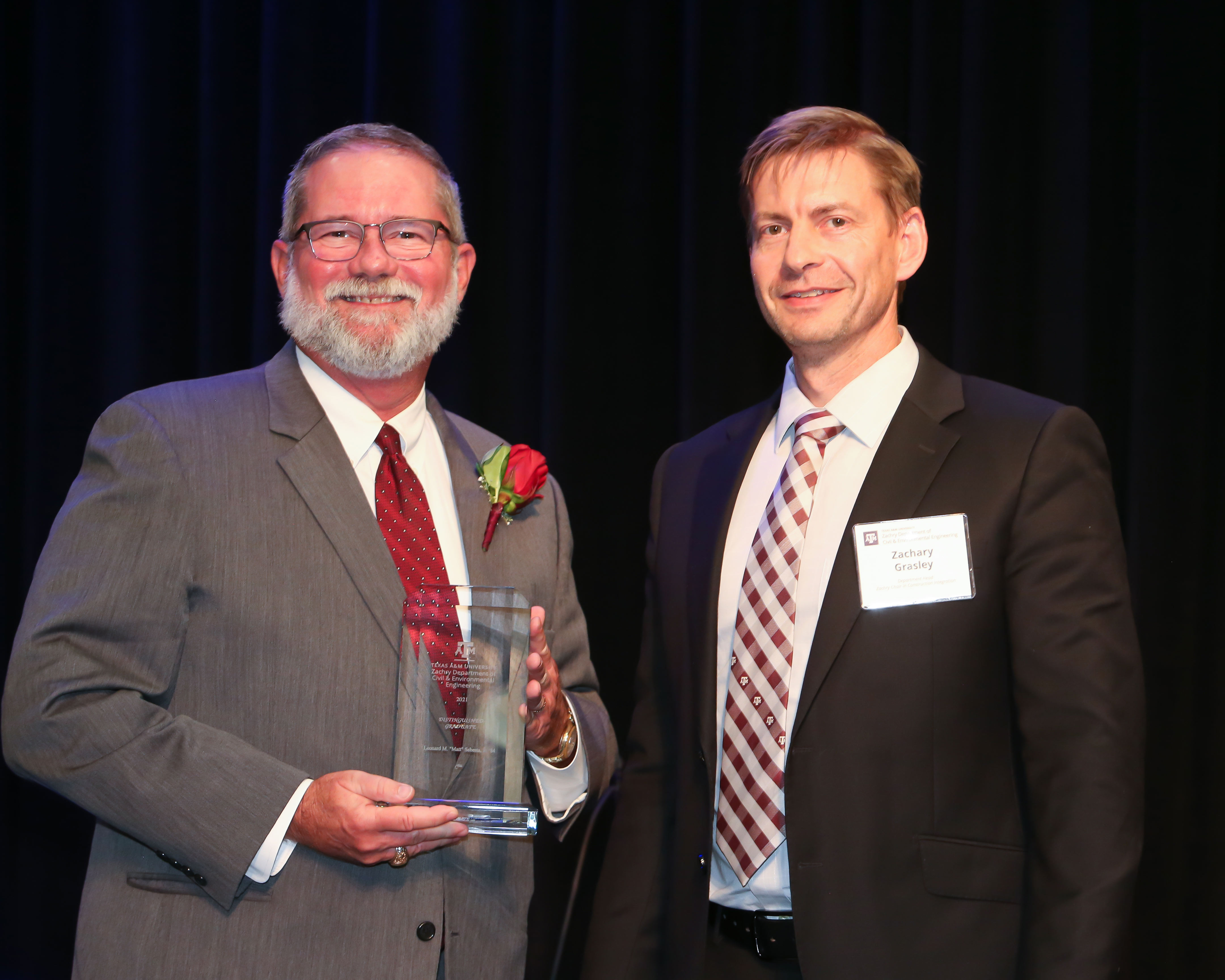 Man dressed in a gray suit with a red tie and rose boutonniere, holds a clear glass award plaque and poses on stage with another man in a dark suit, white shirt, and striped tie.