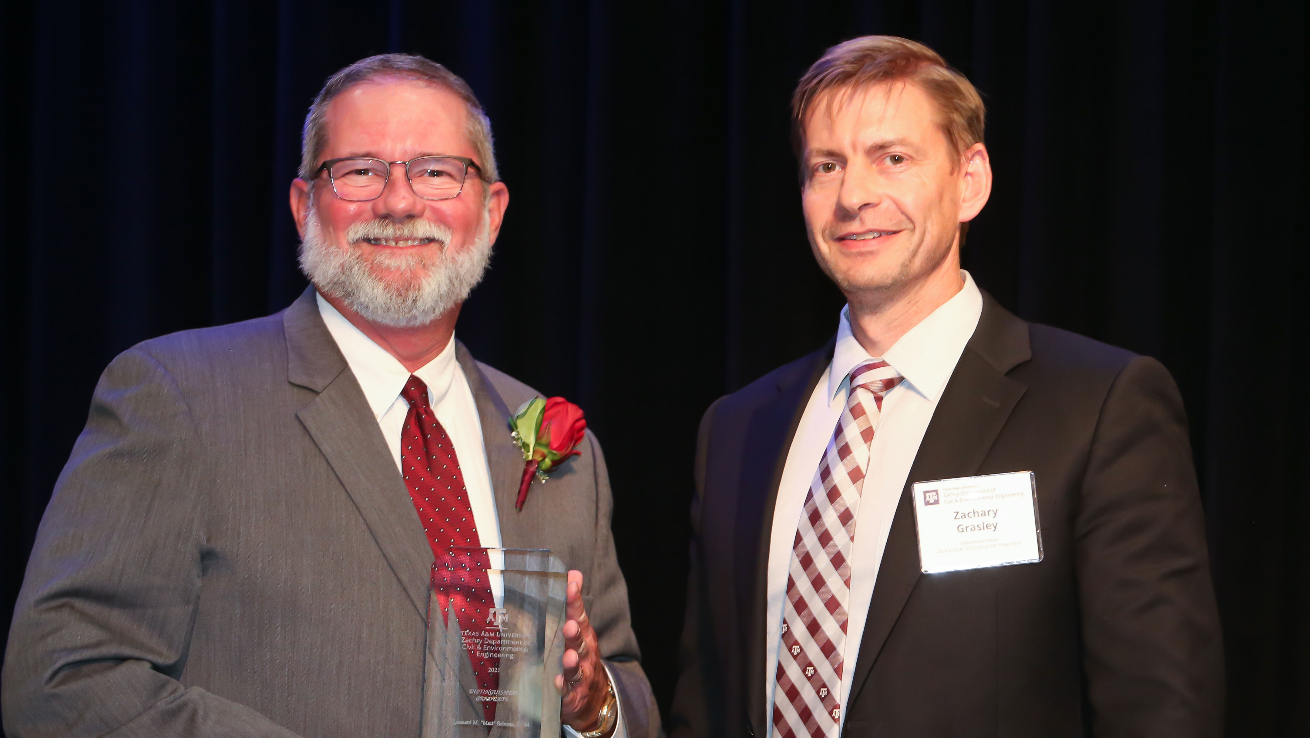 Man dressed in a gray suit with a red tie and rose boutonniere, holds a clear glass award plaque and poses on stage with another man in a dark suit, white shirt, and striped tie. 