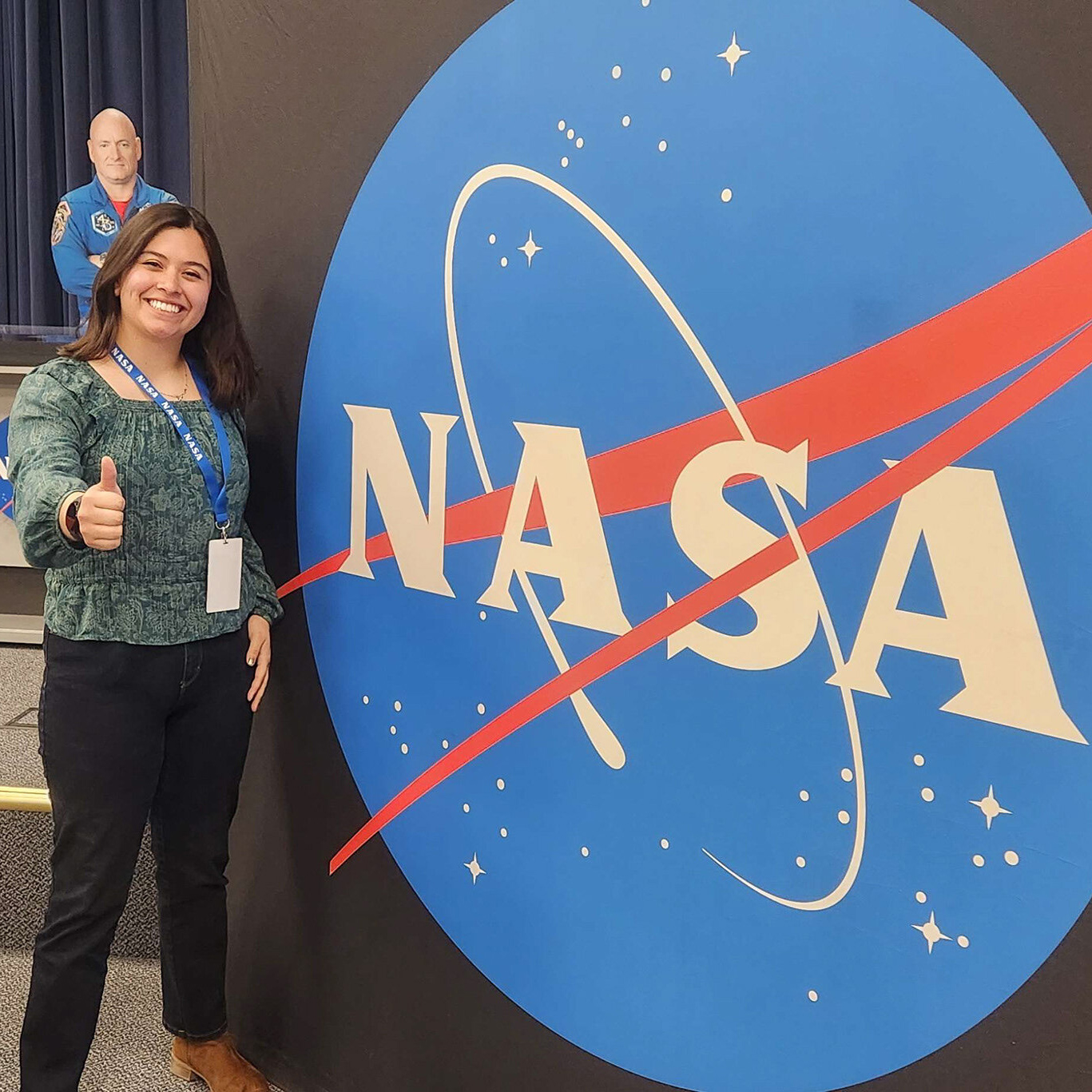 A woman wears a teal blouse and smiles with a thumbs up next to a large sign with the NASA logo printed. 