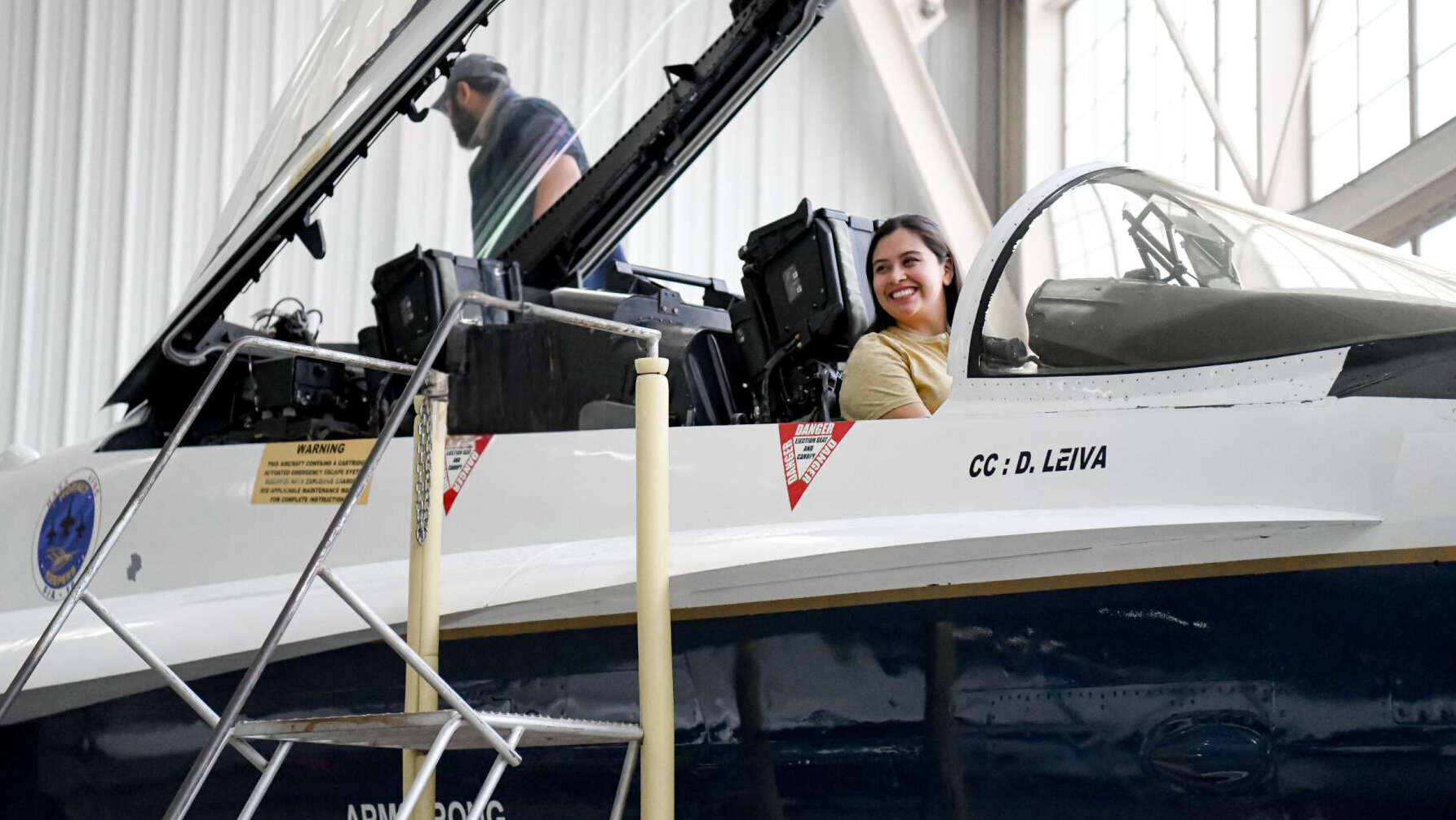  A woman smiles in a yellow t-shirt as she sits in a white aircraft in an airplane hanger. There is a ladder leading up to the aircraft and a man standing in the distance.
