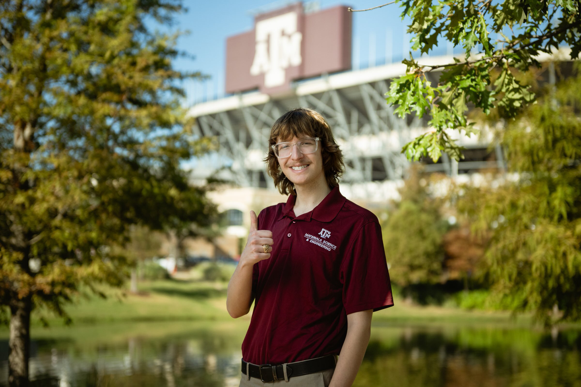 A man smiles and gives a thumbs-up with university football field in the background. 