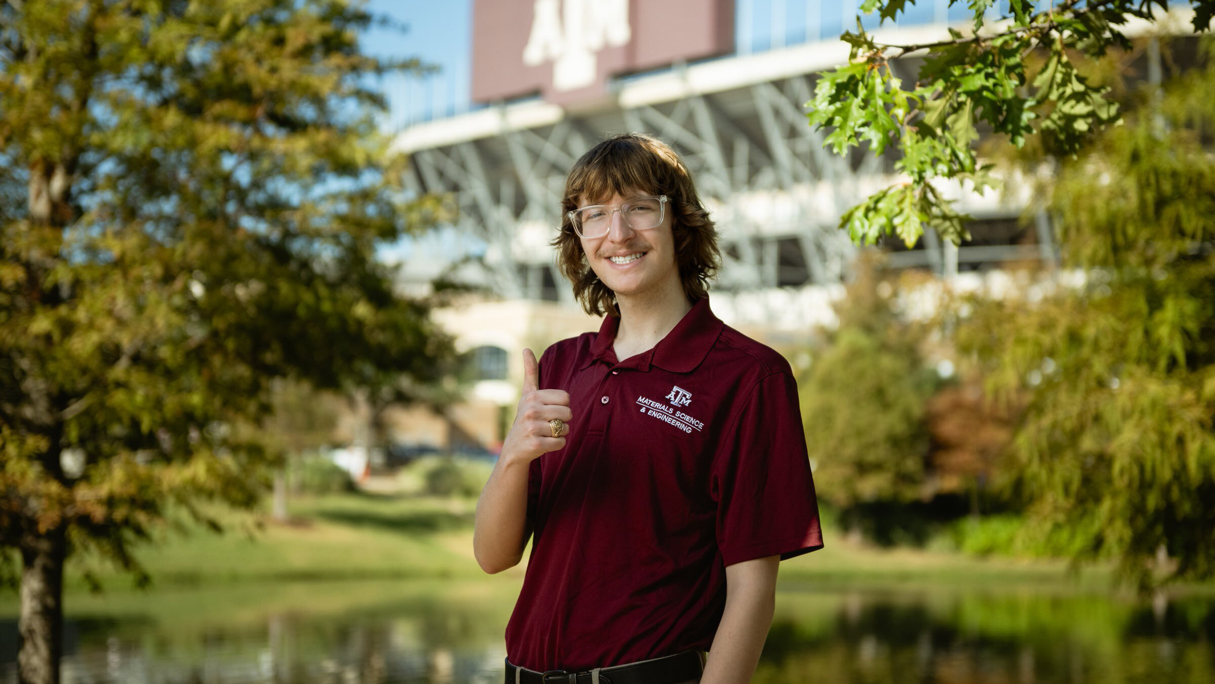 A man smiles and gives a thumbs-up with university football field in the background. 