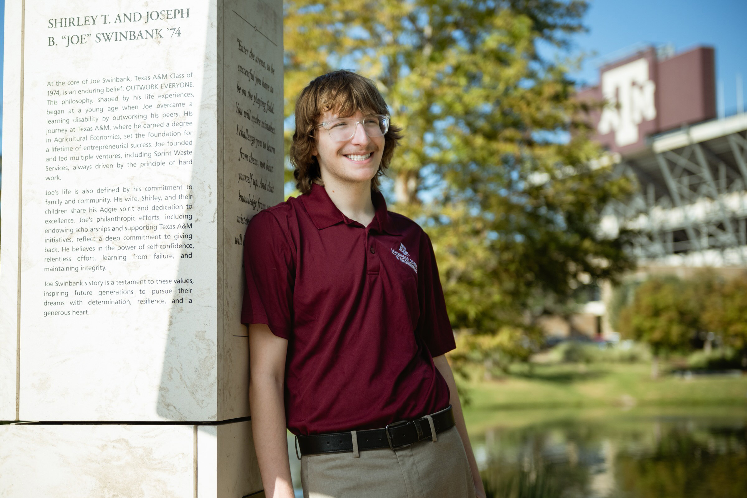 A man leans against the family monument near the university football field on a sunny day. 
