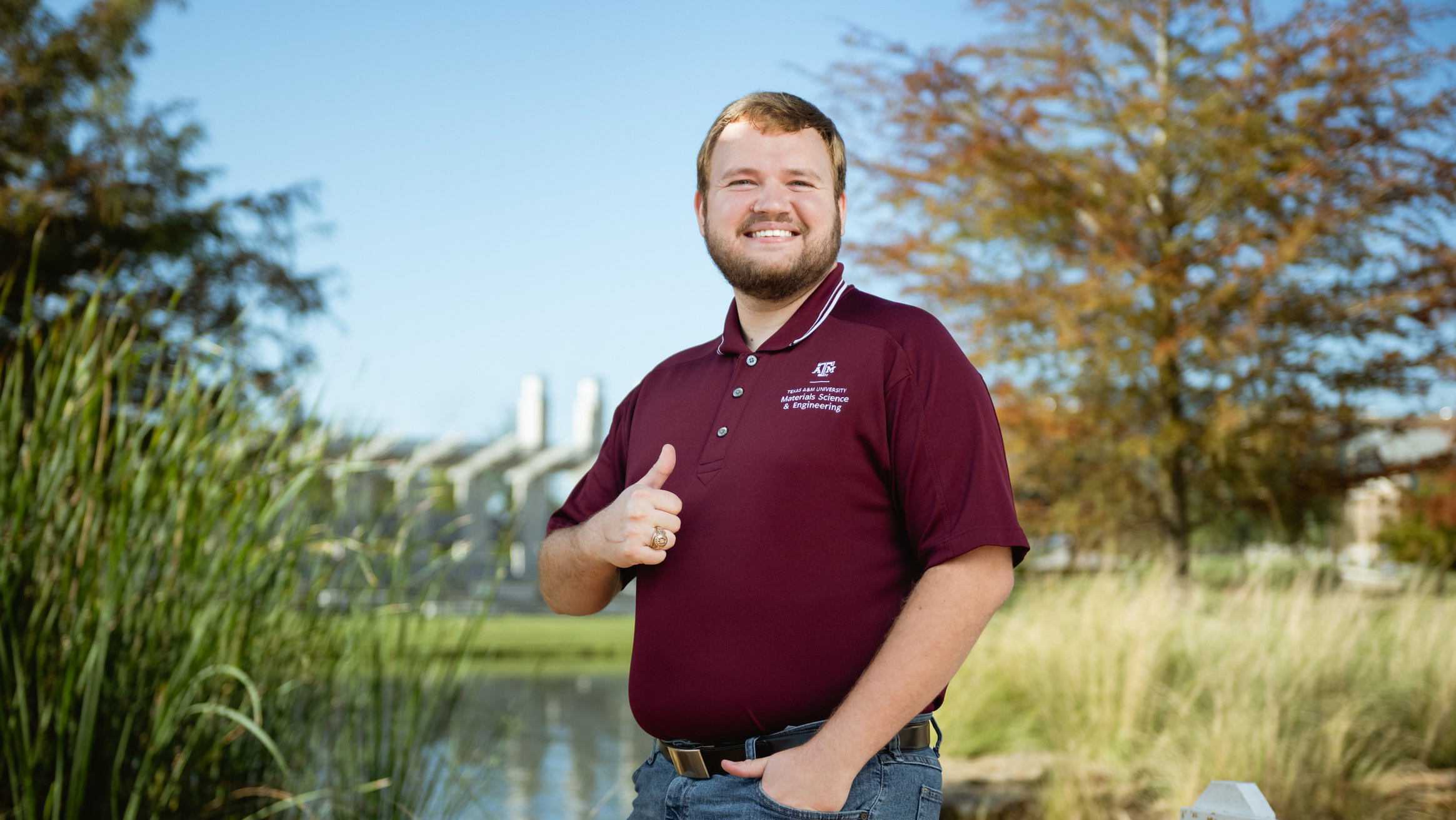 A man smiles and gives a thumbs-up near a pond.
