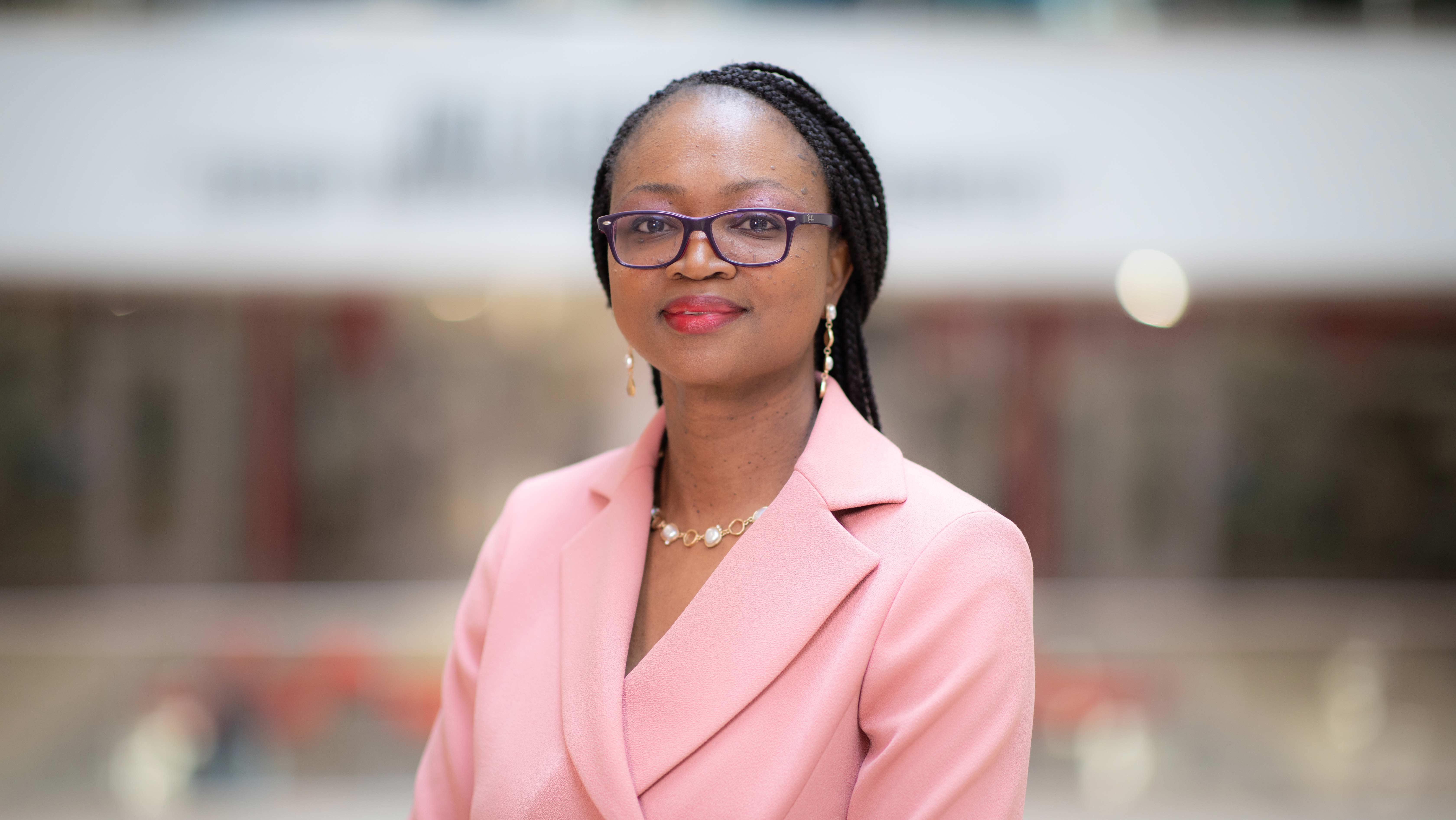  A woman in a pink blazer stands smiling in front of a blurred background. 