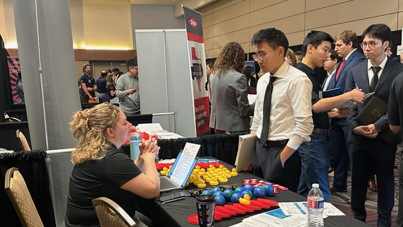 A woman sitting at a booth with a laptop and promotional materials engages in conversation with a man in formal attire. Several people are visible in the background, interacting with other booths at an event venue.