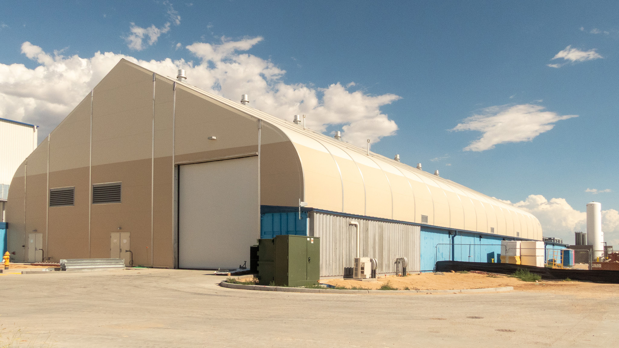 Large warehouse building with a sloped roof in an open industrial area under a blue sky.