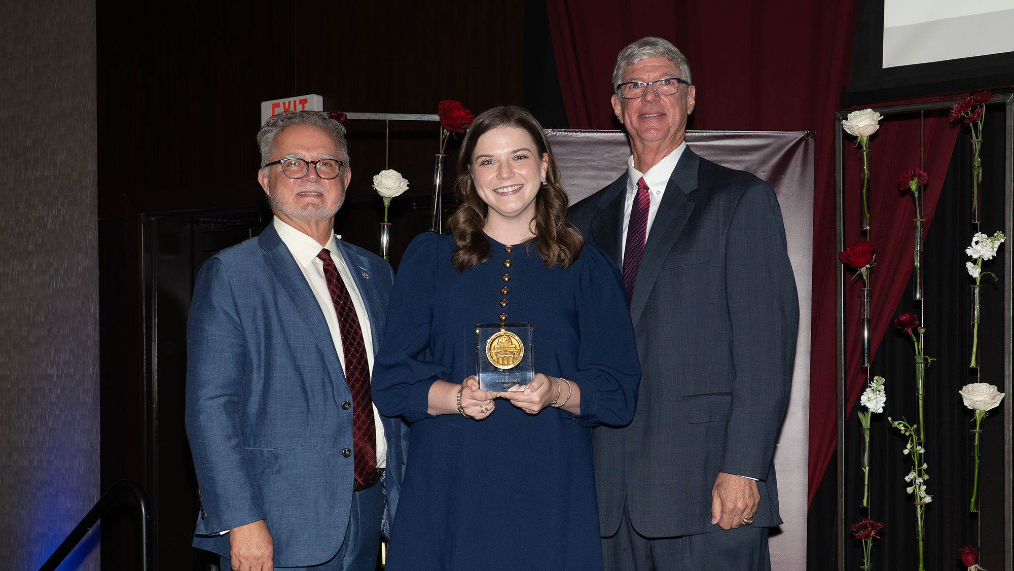 Two individuals stand on a stage, presenting an award to a third person that stands in-between them. 