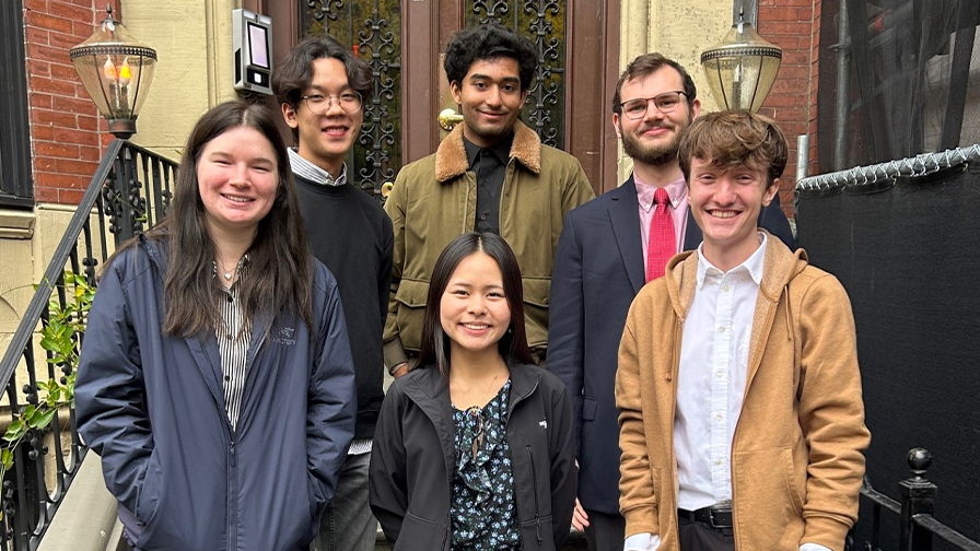 A group of six individuals poses on the steps of a building. They stand in two rows, with three people in the back and three in the front.  
