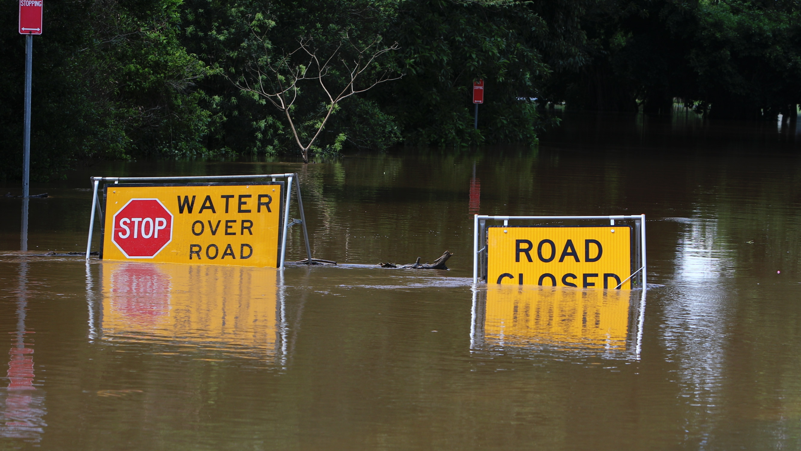 A flooded road showing two signs half covered with water that read water over road and road closed.