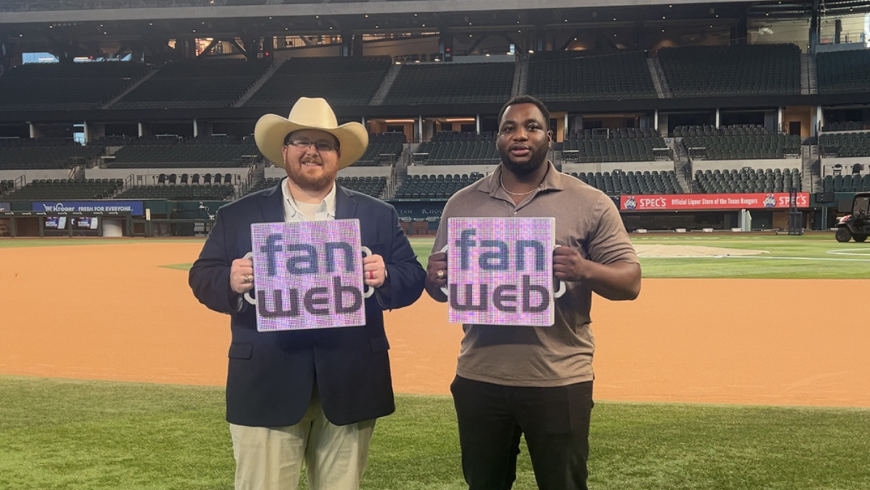 Two men stand on a football field while holding digital tablets that display the word FanWeb.