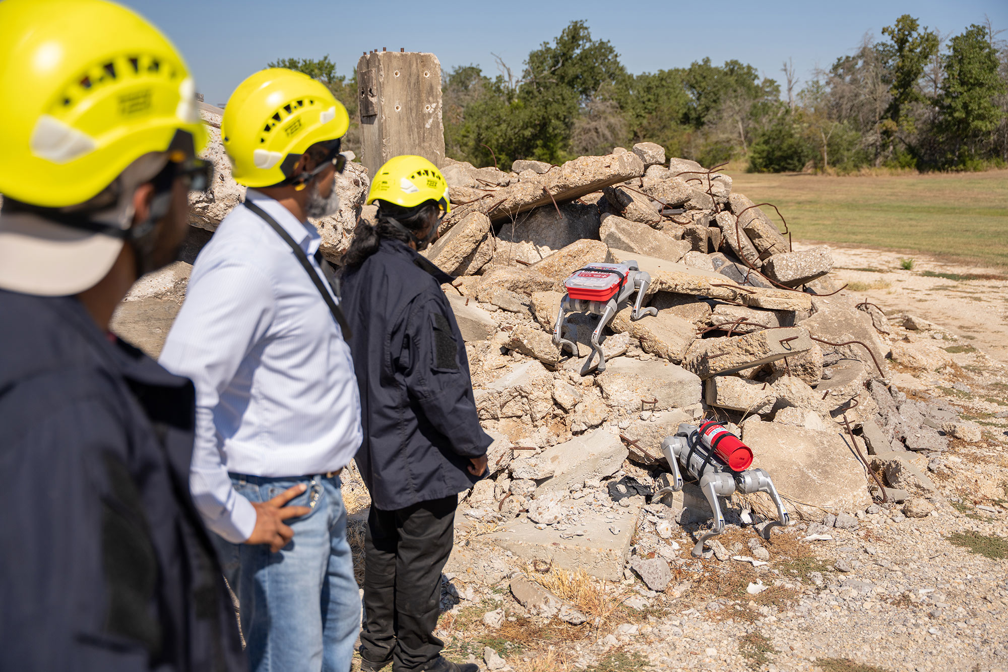 Two four-legged robots climb concrete rubble with a first aid kit and fire extinguisher strapped to their backs while three researchers watch.