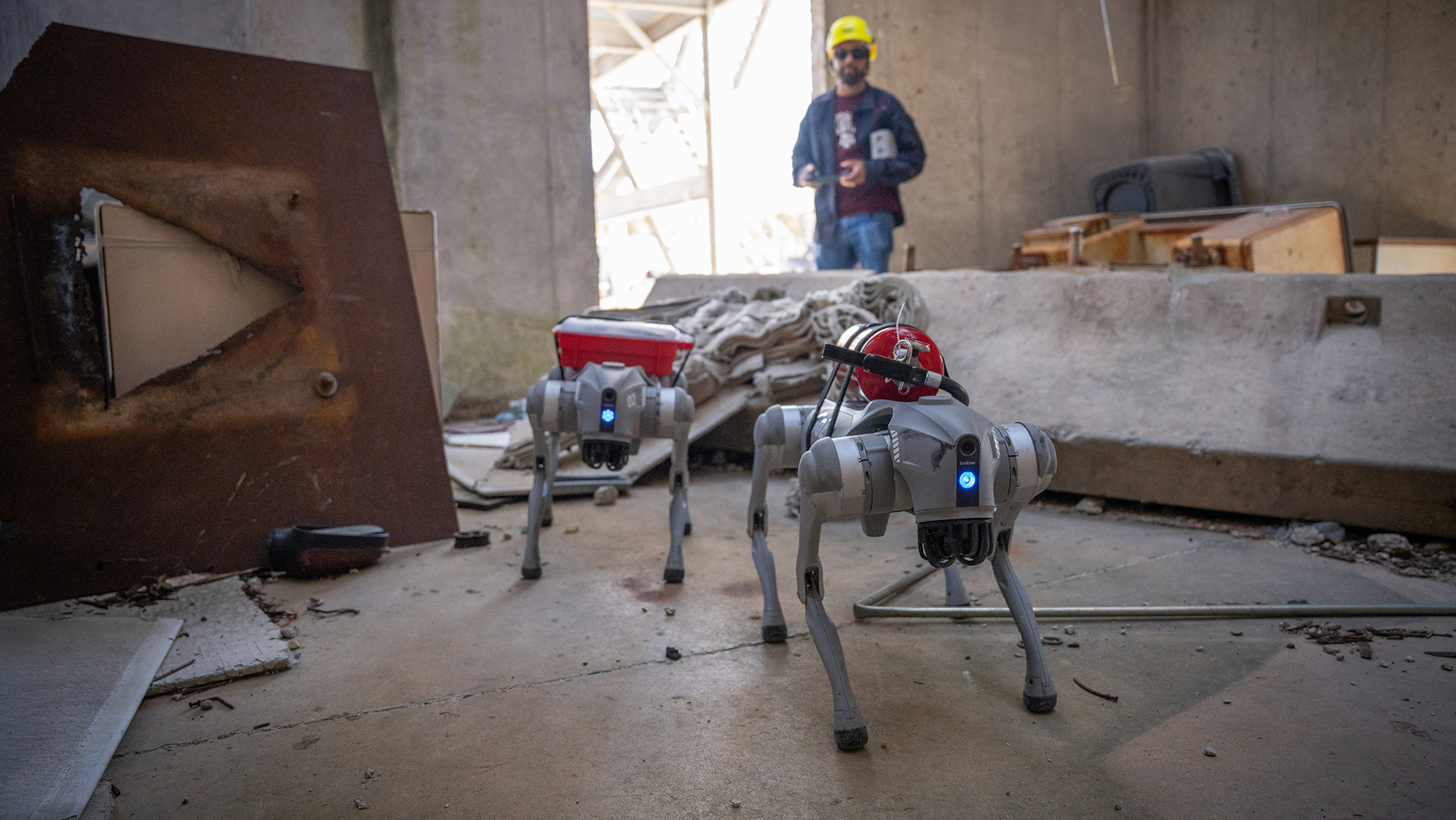 Two four-legged robots walking through rubble with a man behind them.