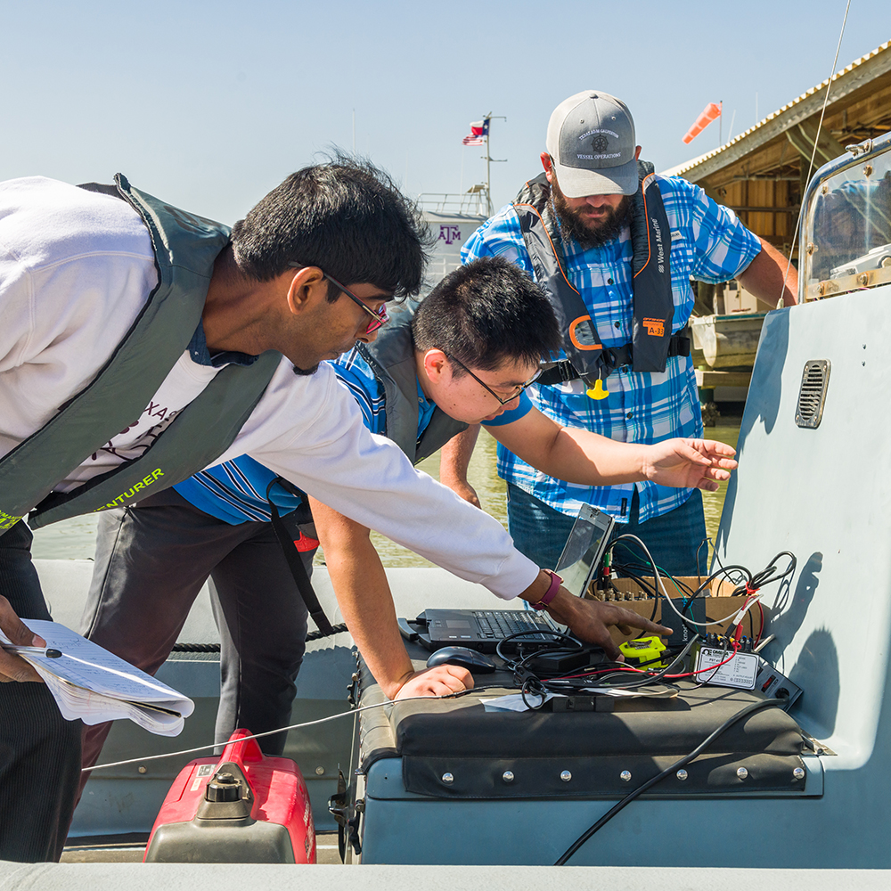 Three researchers on a boat are connecting wiring to a computer laptop and arranging other instruments for an experiment.
