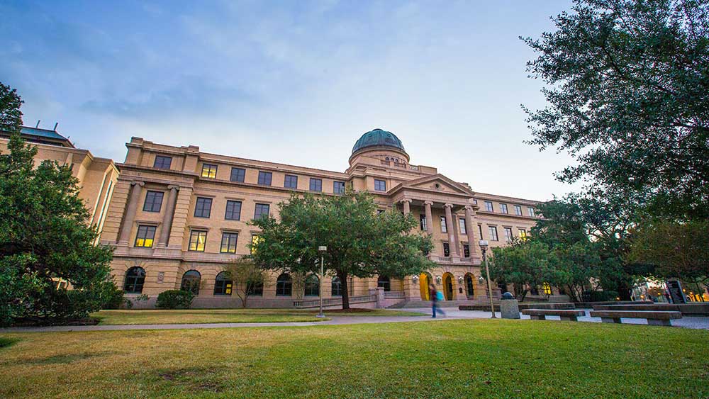 Texas A&amp;M University Academic Building, a 1914 four-story brick and stone structure highlighted with a central column-supported overhang shading a second-story porch and a central copper dome on the roof. 