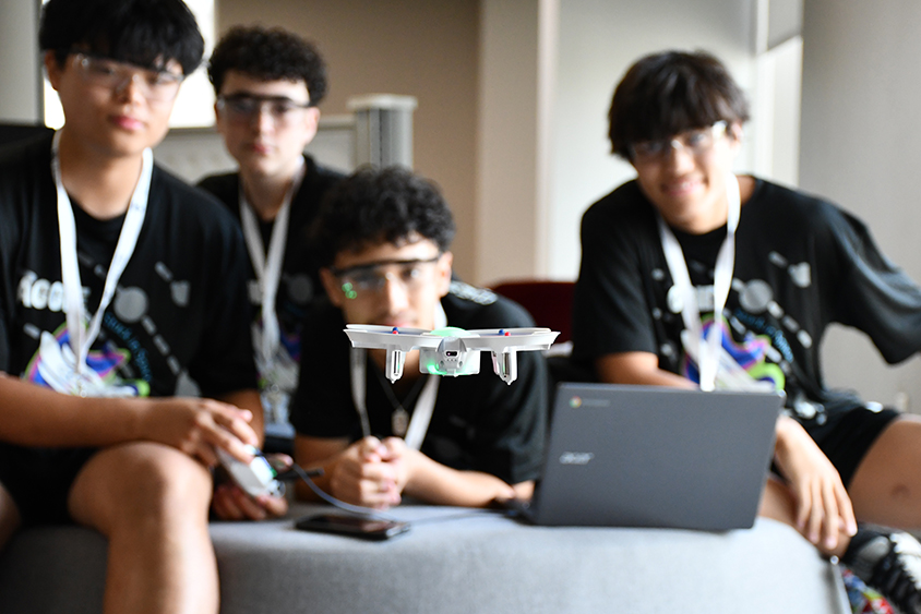 Four male high schoolers wearing black shirts and safety goggles control a small drone that they programmed. 